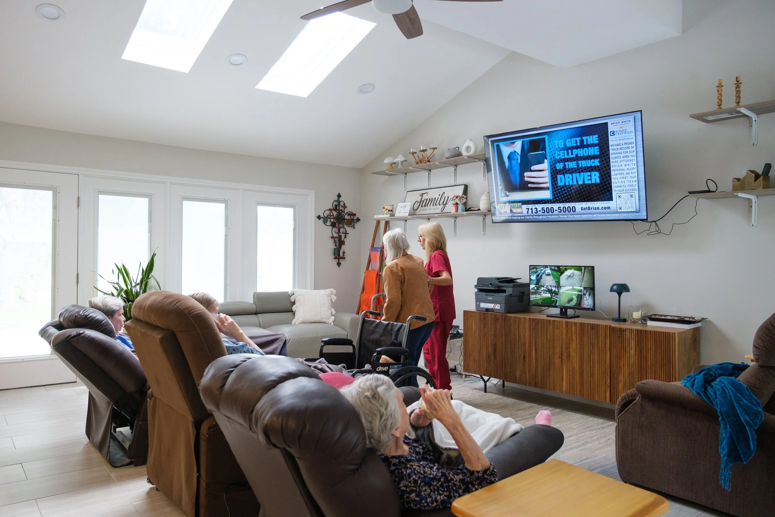A living room with a group of people gathered, including elderly women seated in recliners and a woman in red standing. The room has large windows, a wall-mounted TV displaying an ad, shelves with decorative items, and a ceiling with skylights and a 