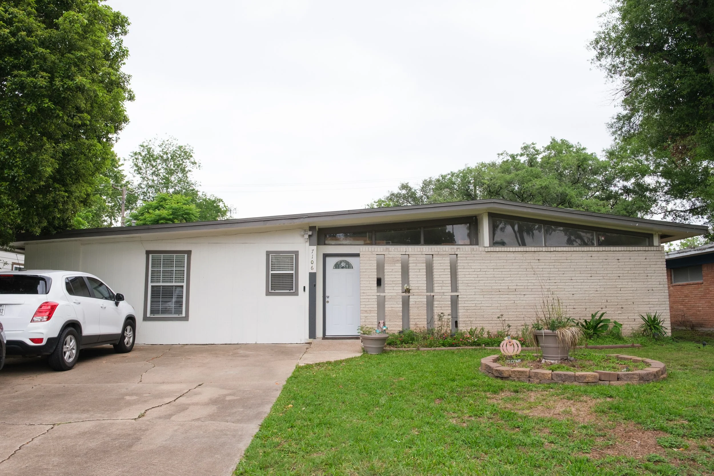 A single-story house with a modern design, white exterior walls, and a flat roof. There are large windows, a front door with a decorative glass panel, and a small front yard with a lawn, flower pots, and garden decorations. Cars are parked in the dri
