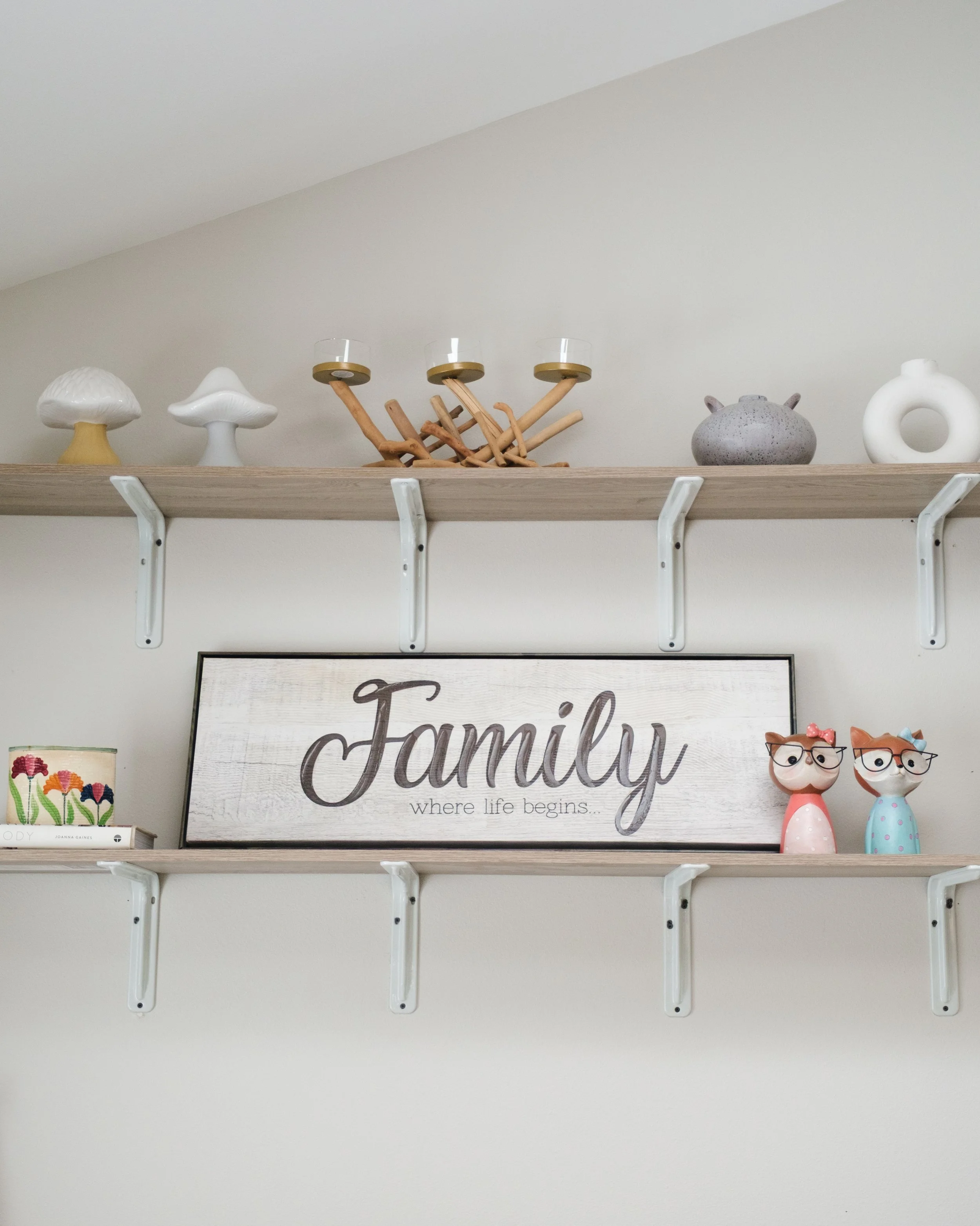 Decorative shelf with mushroom figurines, a candle holder with candles, a framed sign that says 'Family where life begins,' and two small cat figurines with glasses and bows.