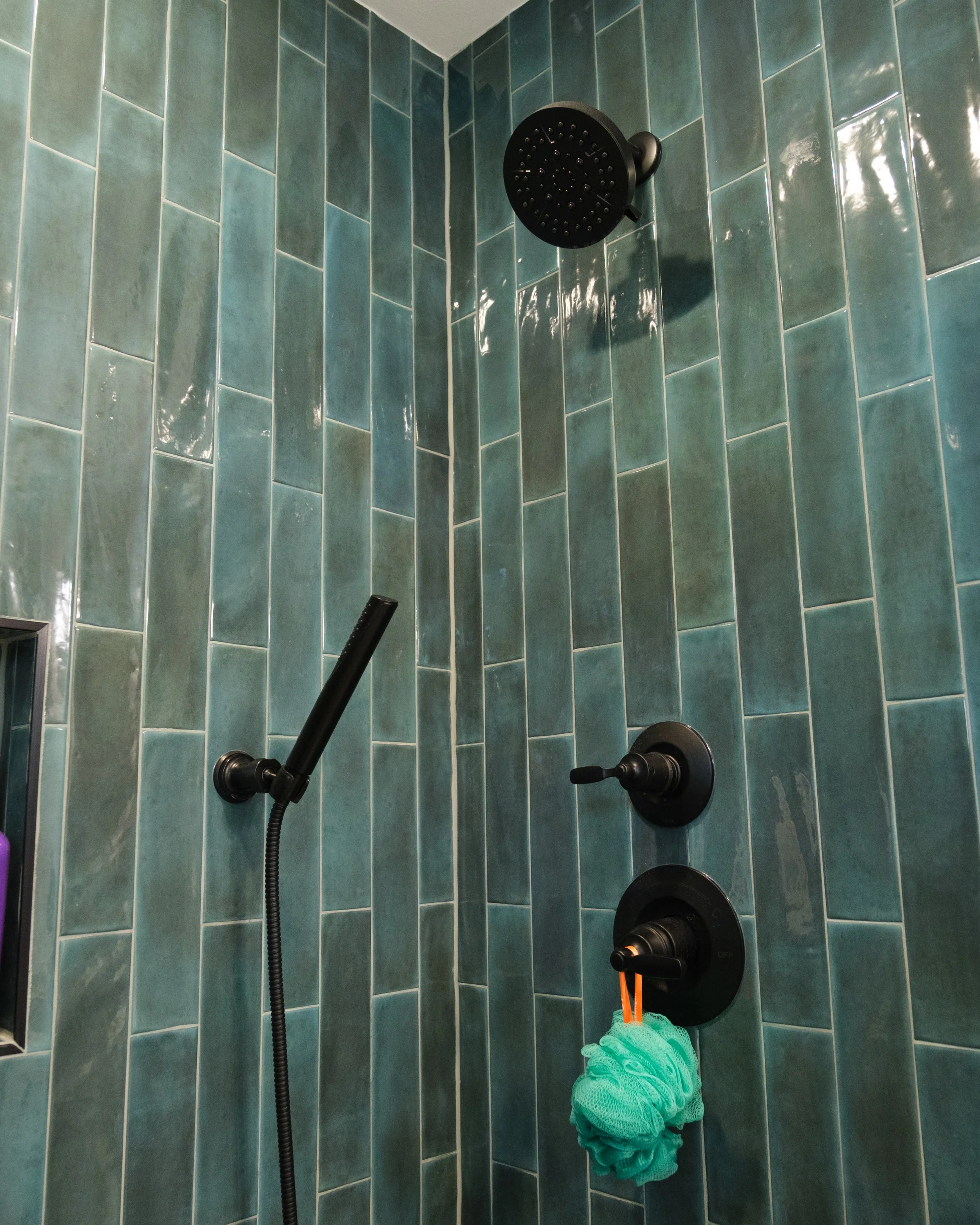 A shower with green tile walls, a black showerhead, a black valve, a black handle, and a green bath sponge hanging from an orange hook.
