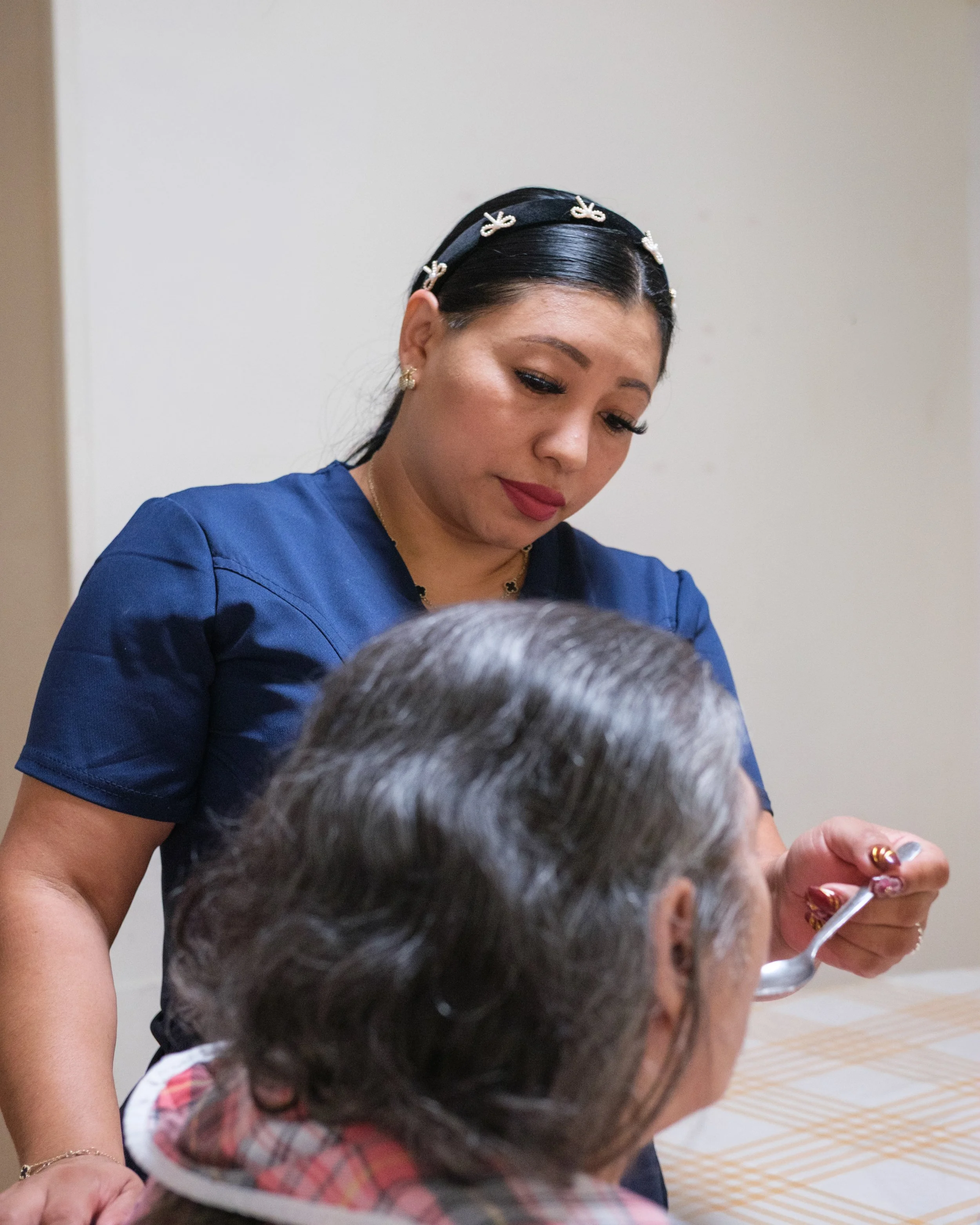 A woman with dark hair and a black headband with white accents is giving a vaccine shot to an elderly person. The woman is wearing a blue top and earrings, focusing on the procedure.