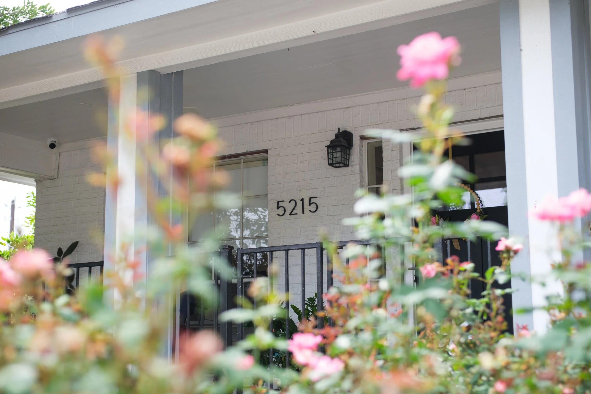 Close-up of pink flowers in front of a house with the number 5215, black railing, and a gray and white exterior with a lantern-style outdoor light fixture.