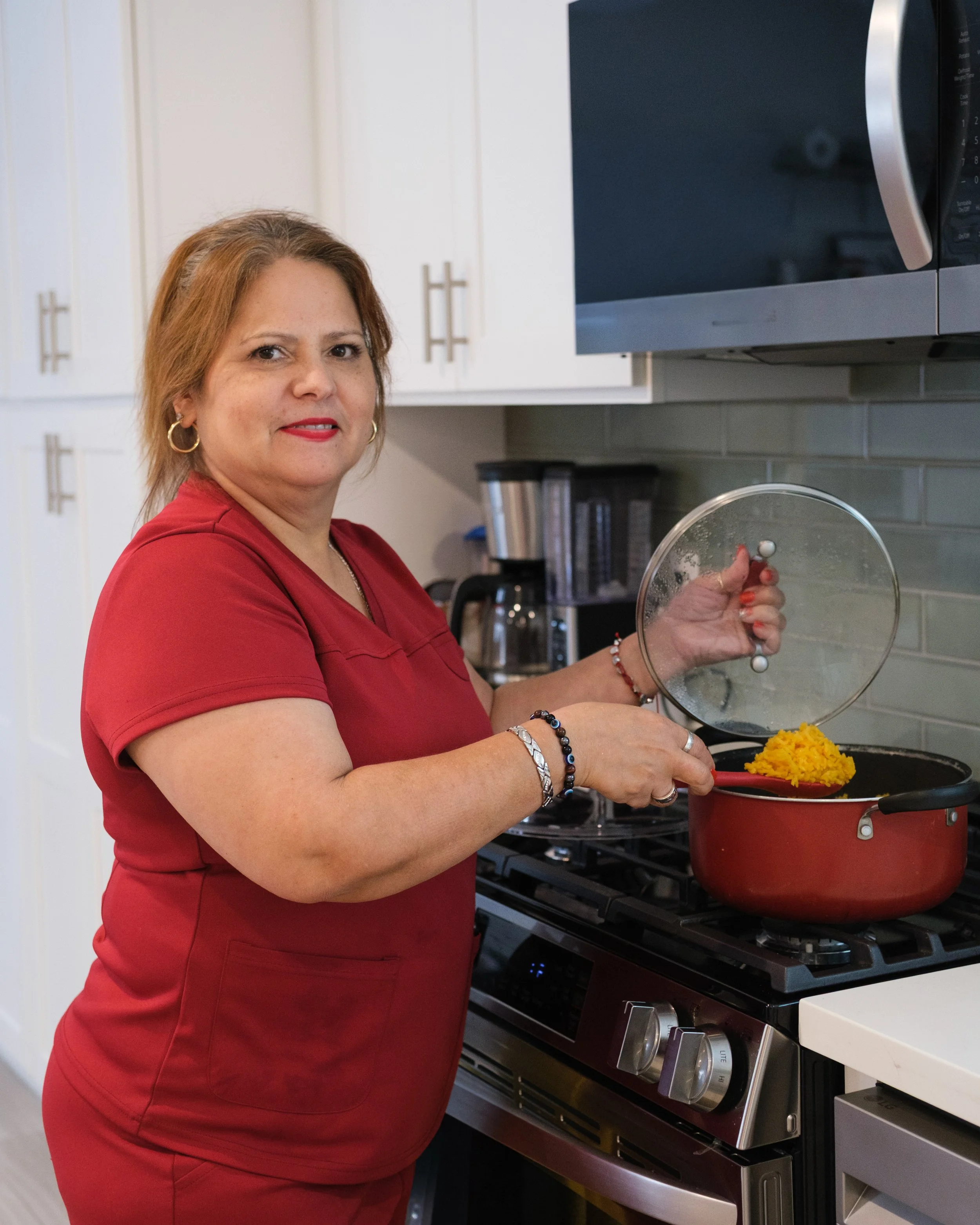 A woman with light brown hair wearing a red shirt stands in a kitchen, cooking on a stove. She is holding a glass lid in her left hand and a red spoon in her right hand, stirring yellow rice in a red pot. There are white cabinets with silver handles 