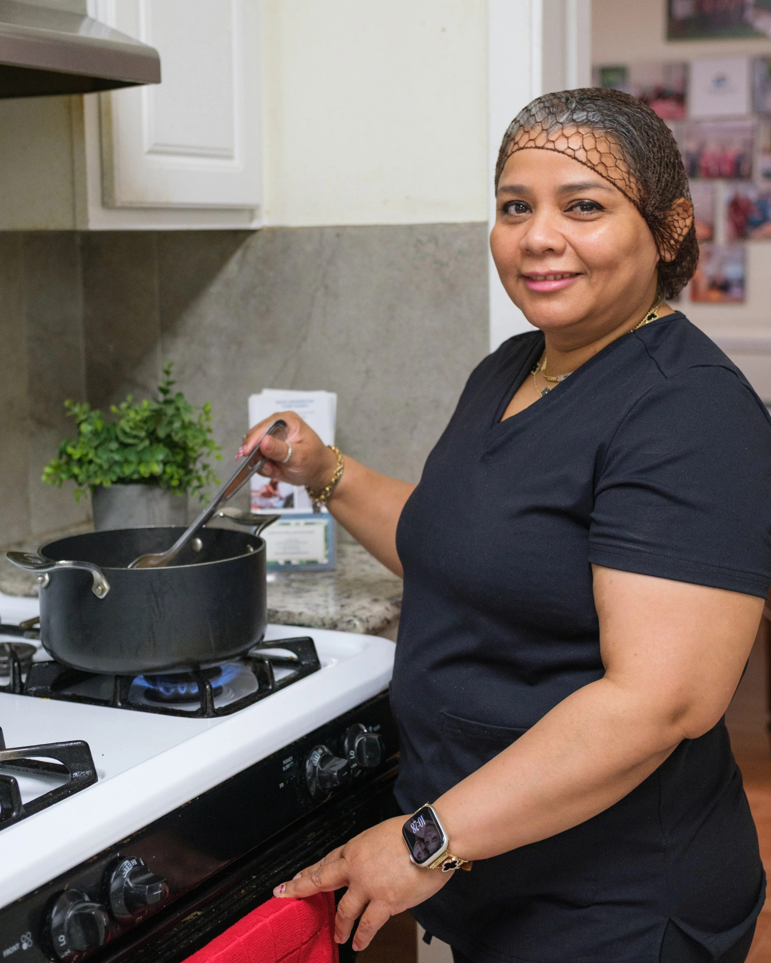 A woman wearing a black shirt, a fishnet hair cover, and jewelry is cooking on a stove. She has a smile on her face and is holding a spoon in a pot on the stove. There are plants and various kitchen items in the background.