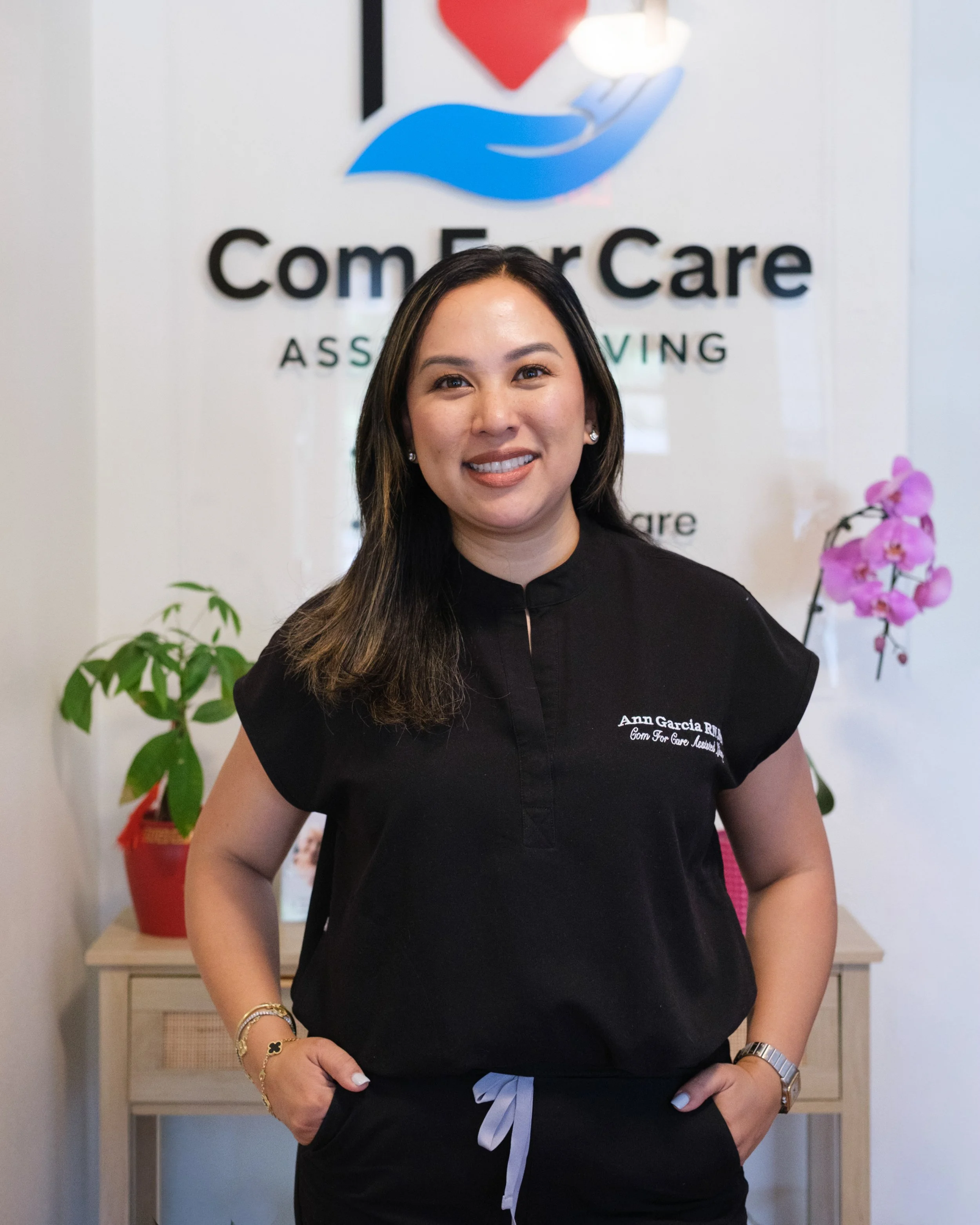 A woman with long dark hair, wearing a black top with a white tie at the waist, and accessorized with jewelry, standing indoors in front of a sign that reads 'Come For Care.' A potted plant with green leaves and purple flowers is partially visible to her left.