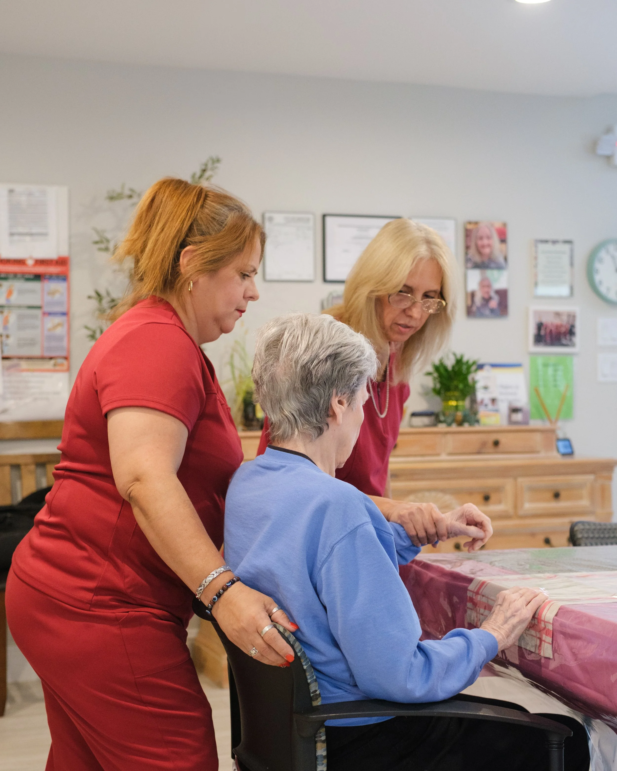Two women assisting an elderly woman seated at a table, in a room with framed documents, pictures, and a clock on the wall.