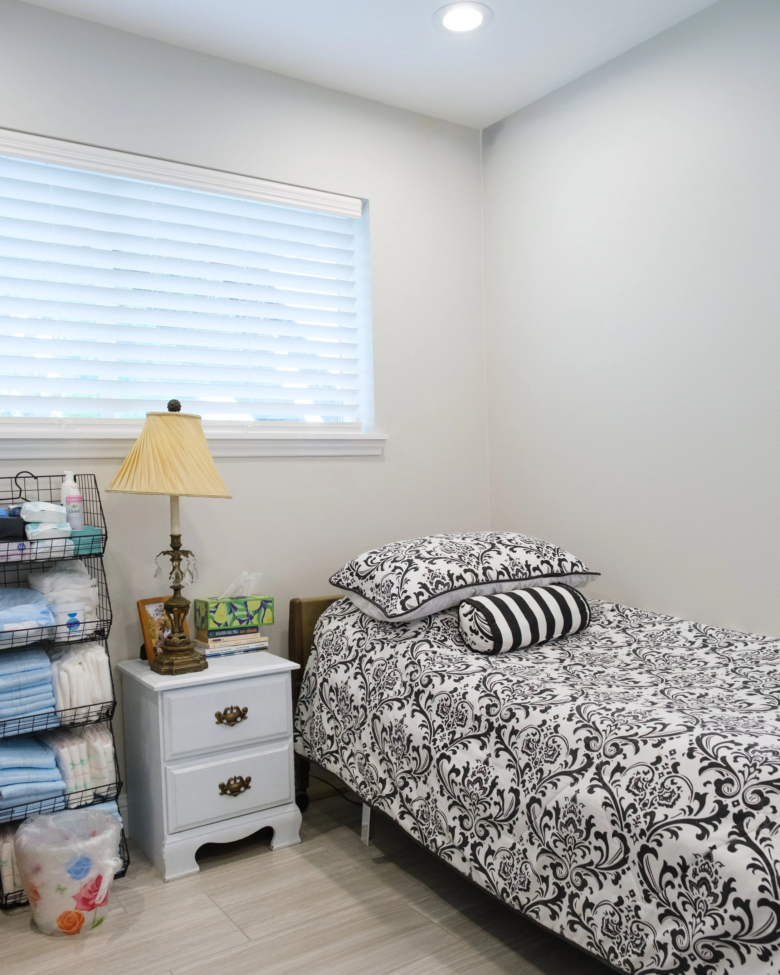 A bedroom with a black and white patterned bed, a white nightstand with three drawers, a vintage-style lamp, a black wire shelf with diapers and toiletries, a window with white blinds, a small framed picture, colorful tissue box, and a trash bin with