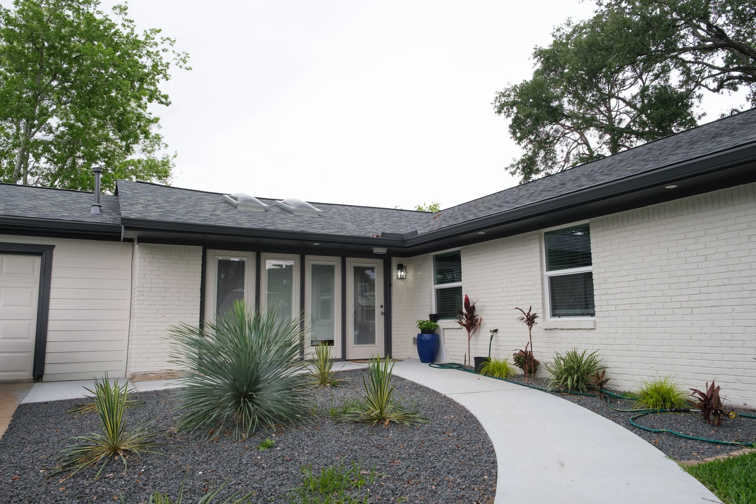Front view of a modern house with white brick exterior, black window trims, a small in-ground patio, and a landscaped garden with various plants and shrubs. There is a concrete walkway leading to the entrance under a covered porch area. Tall trees ar