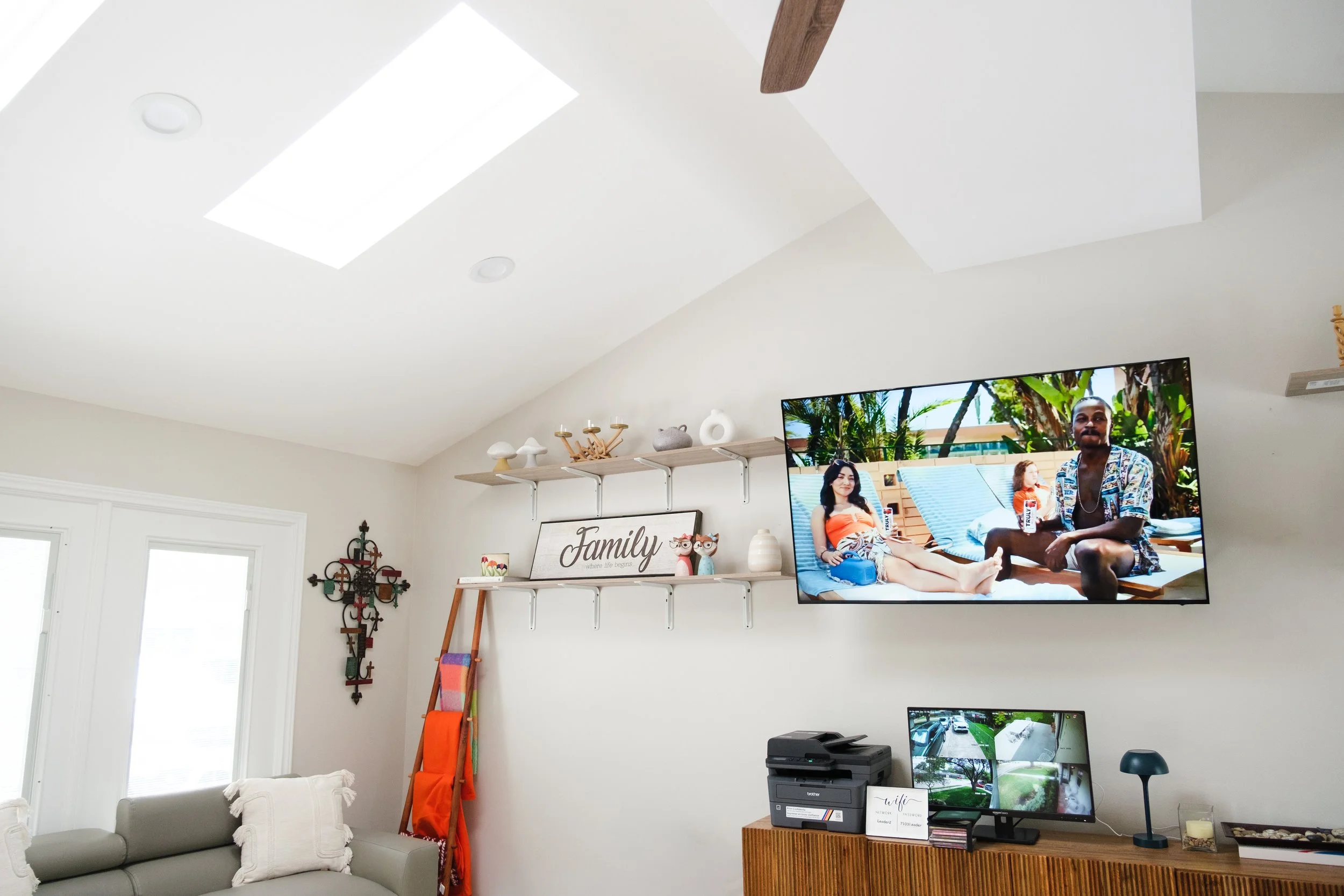 Living room with a high ceiling featuring a skylight and a flat-screen TV displaying a tropical scene. Shelves with decor items, a sign reading 'Family,' and a wall hanging are visible. A gray couch with pillows and a small computer desk are also pre