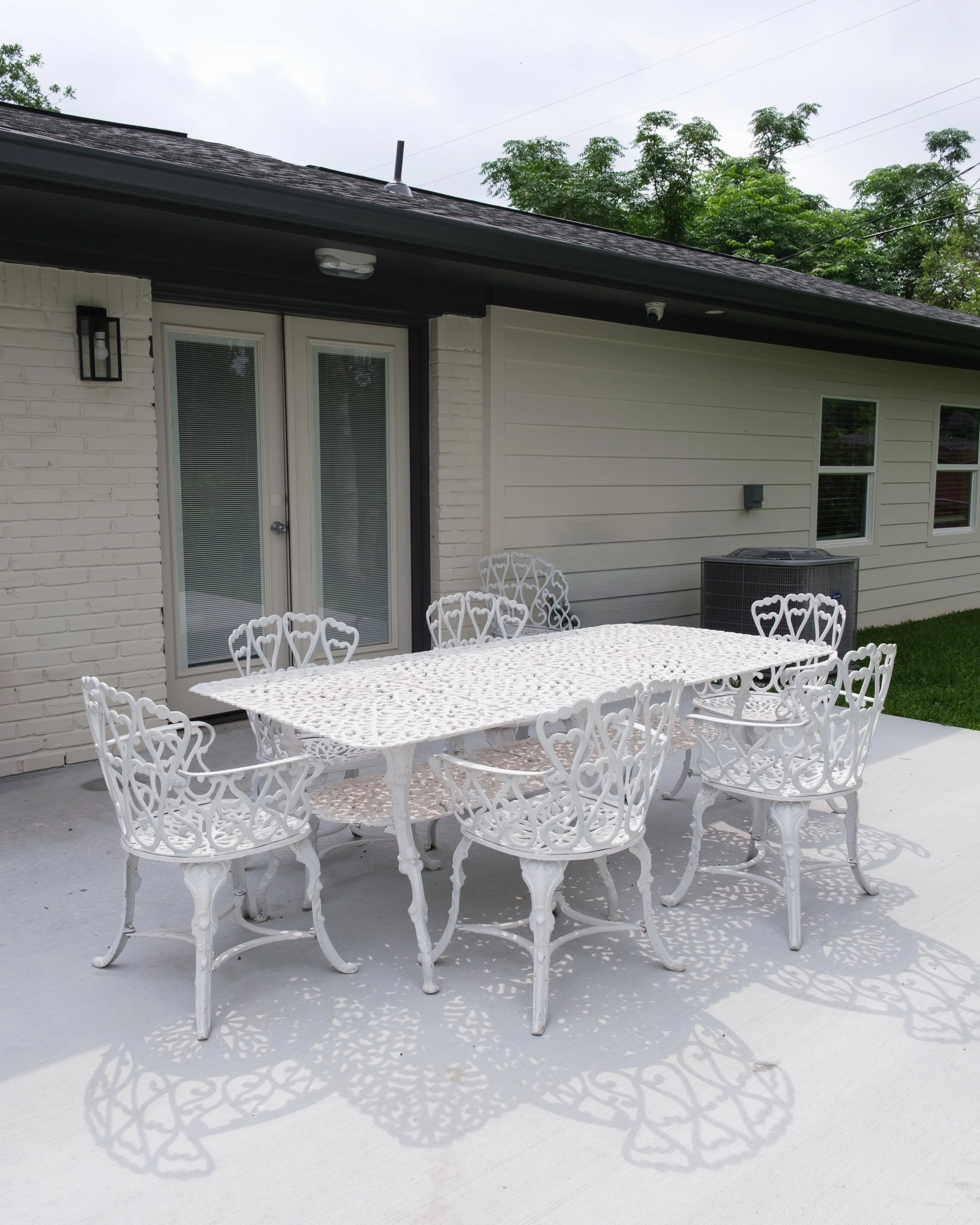 Outdoor patio area with white metal table and matching chairs, house with white siding, glass double doors, windows, outdoor air conditioning unit, and decorative lighting fixture, with trees and power lines in the background.