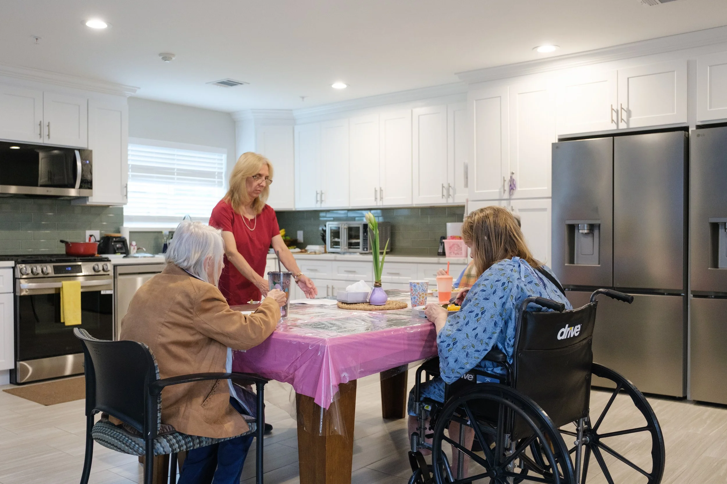 A woman in a red shirt stands at a table in a kitchen, surrounded by three elderly women with disabilities, including one in a wheelchair, seated around a table with pink tablecloth, flowers in a small vase, and drinks. The kitchen has white cabinets