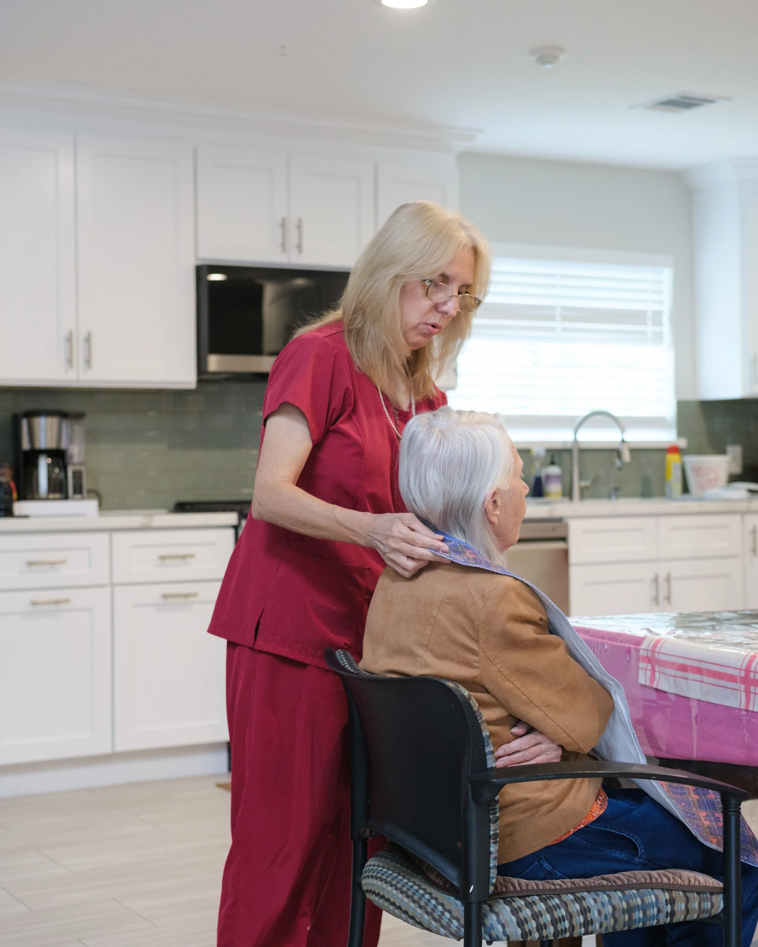 A caregiver with blonde hair and glasses is standing beside an elderly woman with white hair, seated in a wheelchair, in a bright kitchen. The caregiver appears to be providing assistance or care, and the elderly woman is covered with a brown jacket.