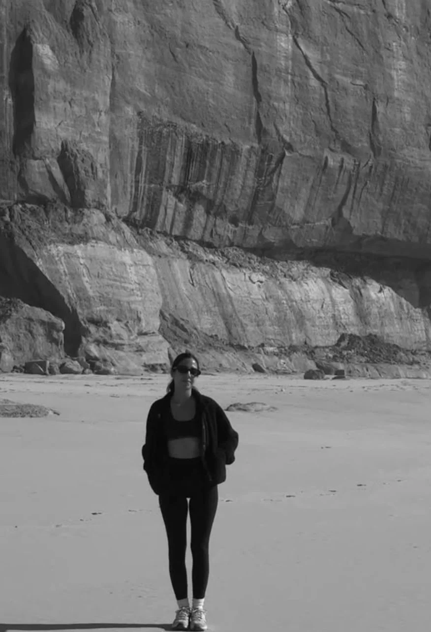 A woman wearing a black jacket, sunglasses, and activewear standing on a sandy beach with large cliffs in the background.