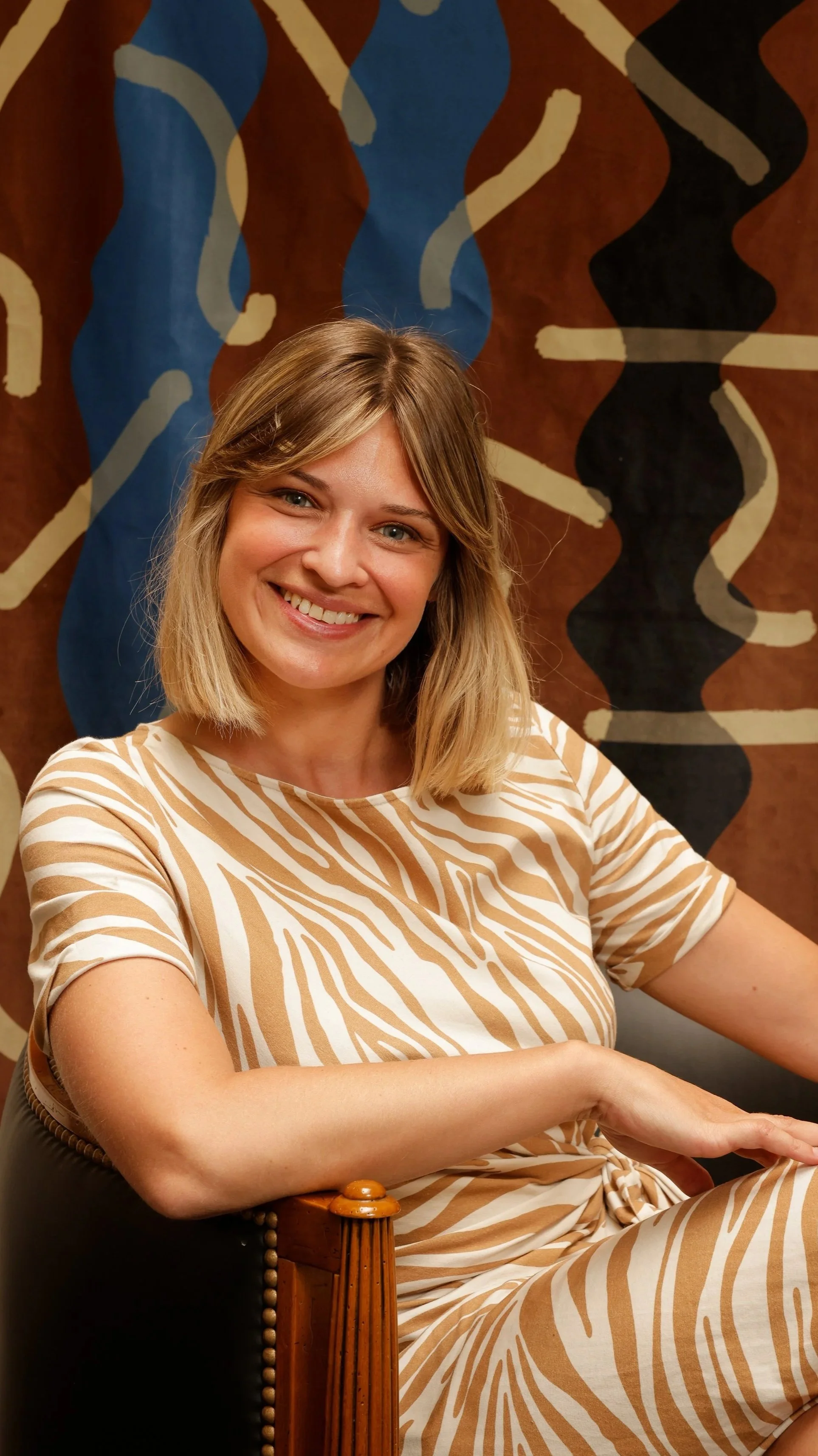 Une femme souriante, aux cheveux blonds, portant une robe rayée beige et blanche devant un fond abstrait avec des formes bleues, noires et beiges. Elle est la fondatrice de l'agence d'architecture d'intérieur Tracé.