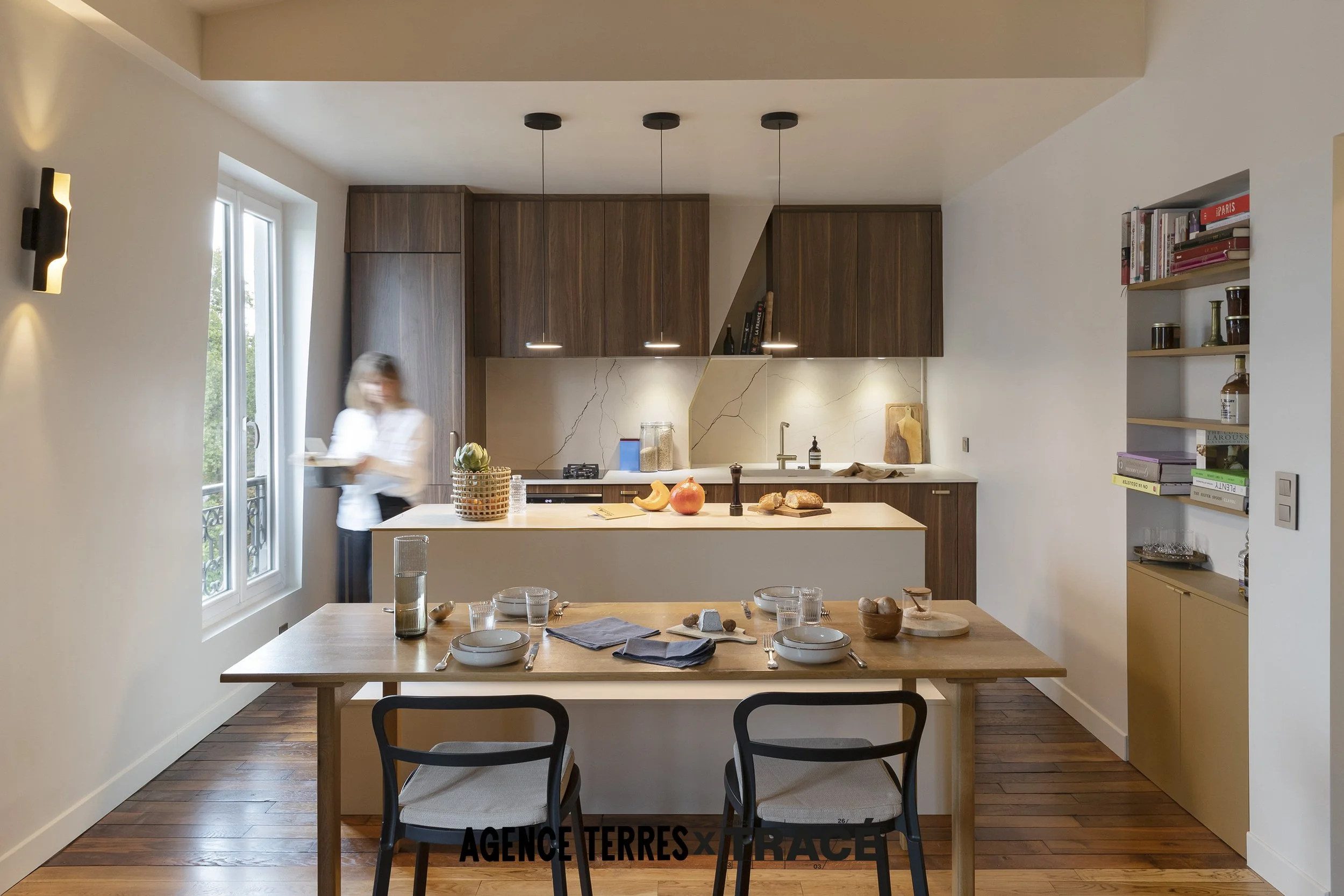Cuisine rénovée avec un ilot central couplé d'une banquette sur mesure pour un coin repas centré dans la pièce. Derrière l'ilot on aperçoit les aménagements sur mesure de la cuisine avec des meubles en bois foncé et une crédence en imitation marbre.