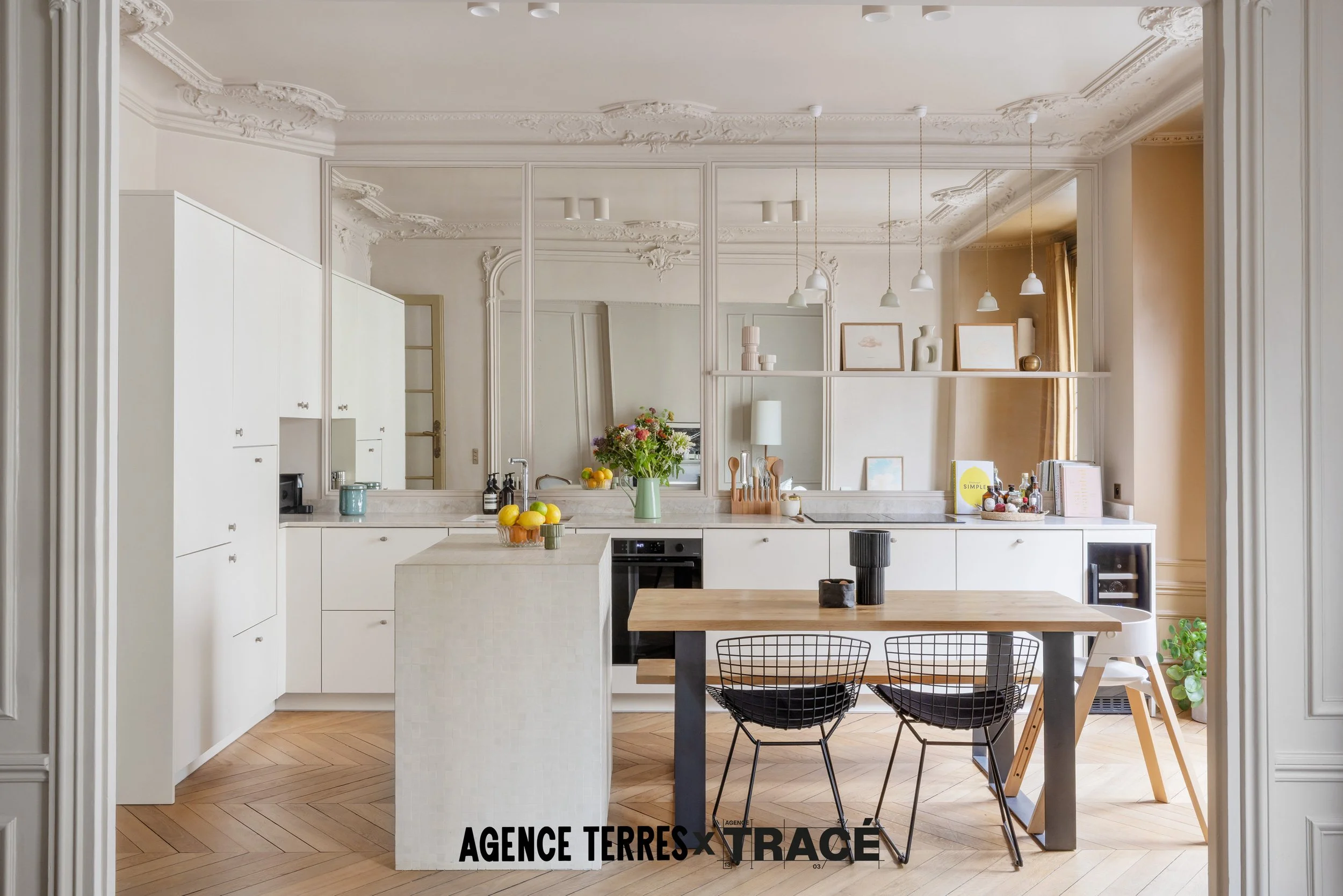 Cuisine ouverte rénovée avec un ilot central carrelé et une table adossée. Linéaire de cuisine sur mesure en arrière plan avec crédence miroir et mur de colonnes de rangements à gauche. Le tout dans une cuisine rénovée claire, lumineuse, chaleureuse.