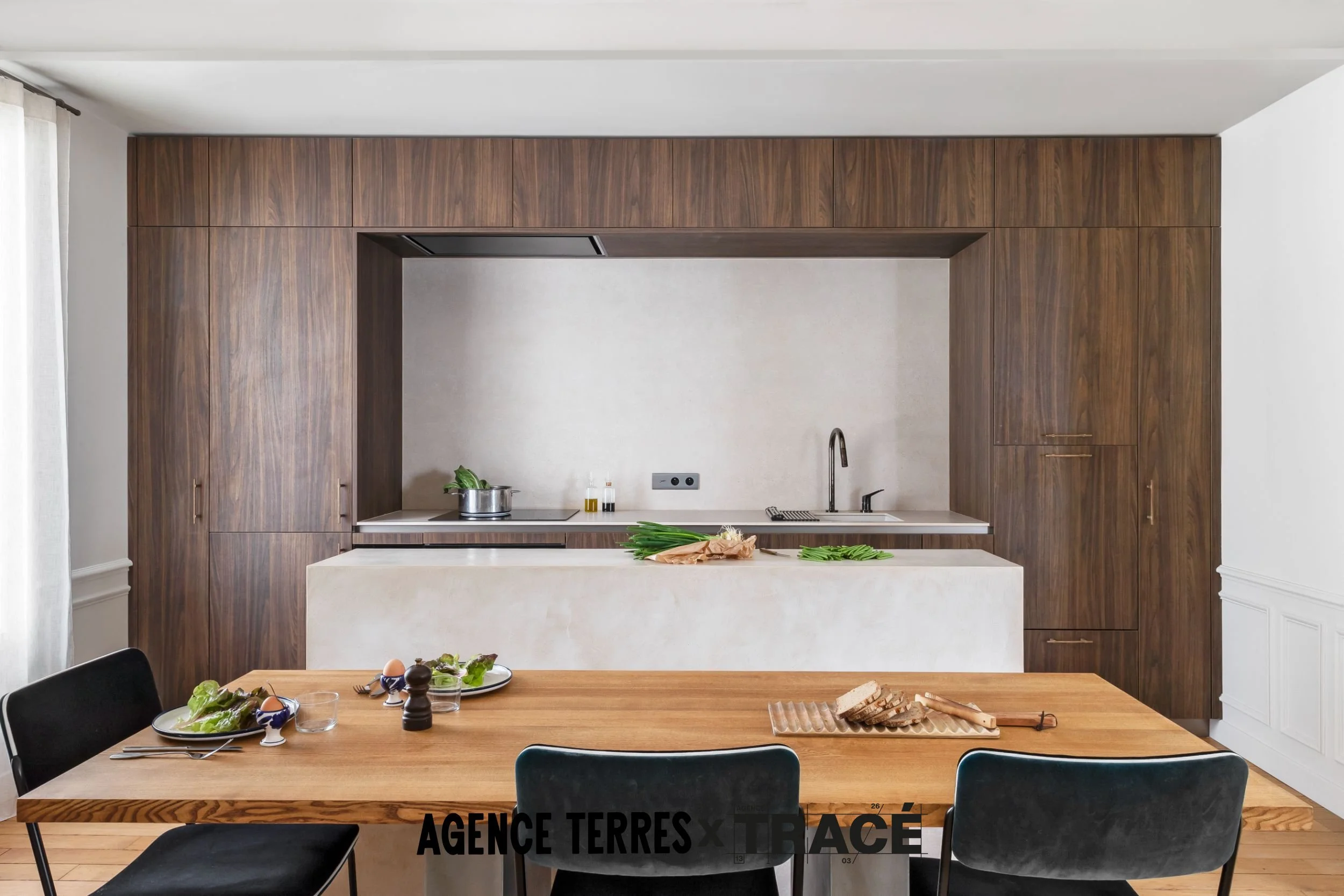 Cuisine sur mesure en noyer et béton ciré beige. La cuisine prend tout l'espace du mur du fond et réalise un cadre de placards sur mesure autour du plan de travail. L'ilot central en béton ciré complète et rajoute une banquette pour la table à manger