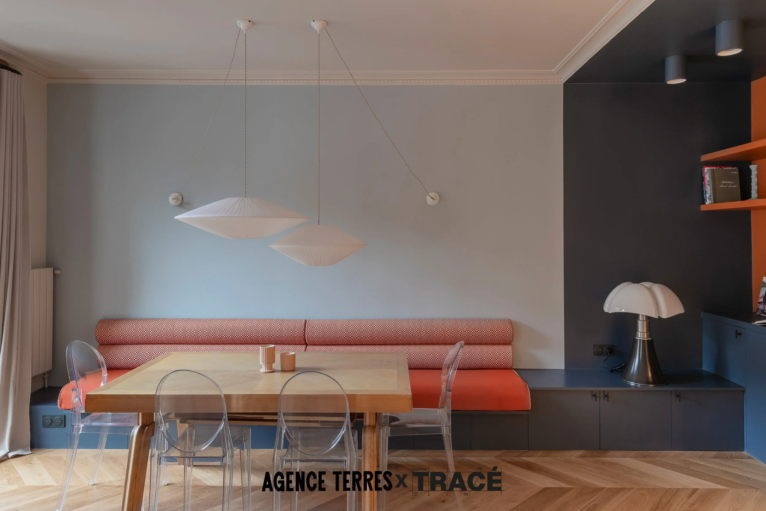 Menuiserie sur mesure avec banquette et coussins sur mesure intégrés pour création d'un coin repas dans un grand salon. Aperçu d'une grande bibliothèque sur mesure à droite, le tout dans les tons orange, bleu foncé et bleu clair.
