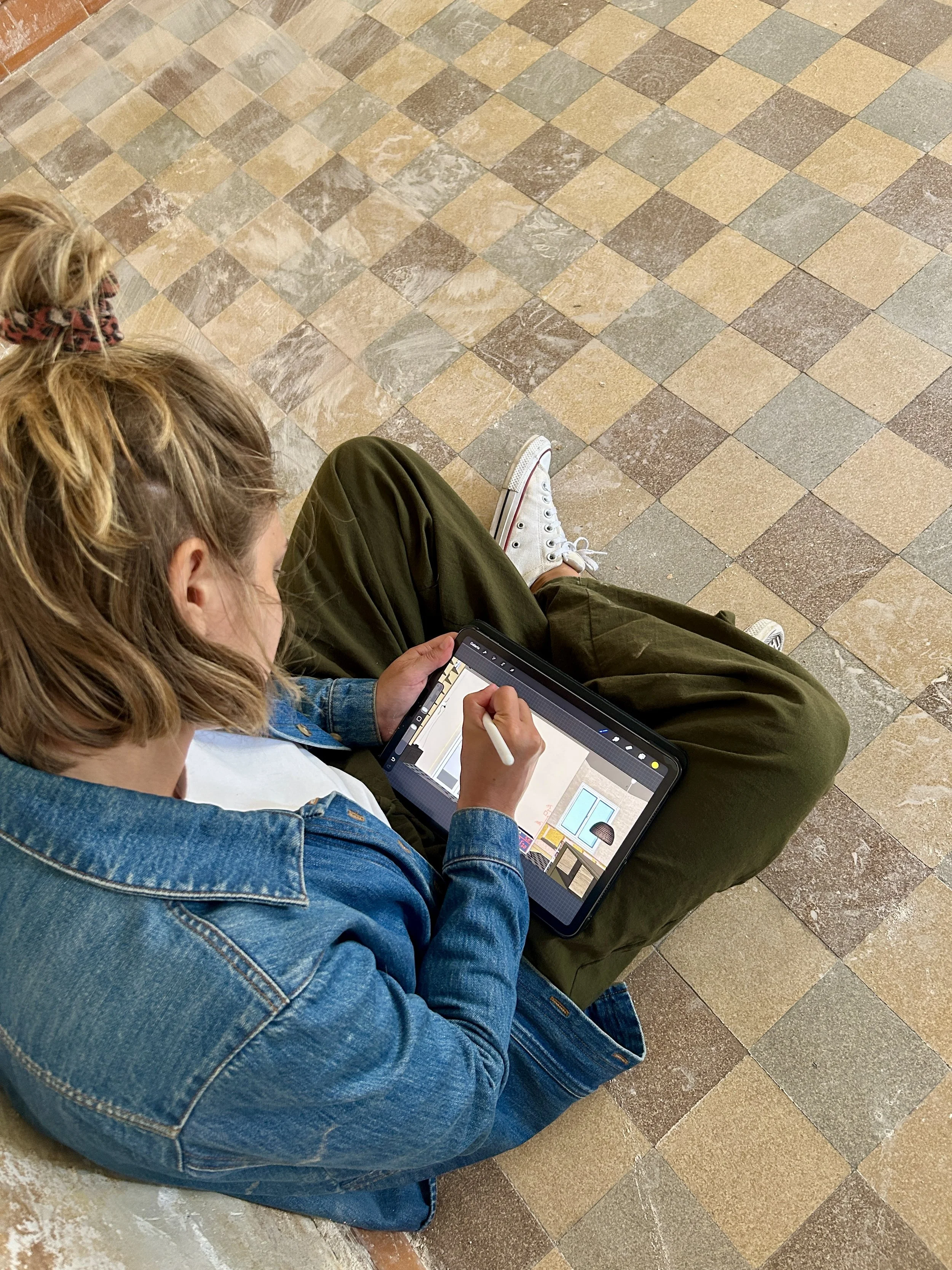 Une personne avec un jean et des baskets blanches assise par terre sur un chantier en rénovation, utilisant une tablette tactile avec un stylet, pour dessiner dessus et annoter des plans d'agencements sur mesure.