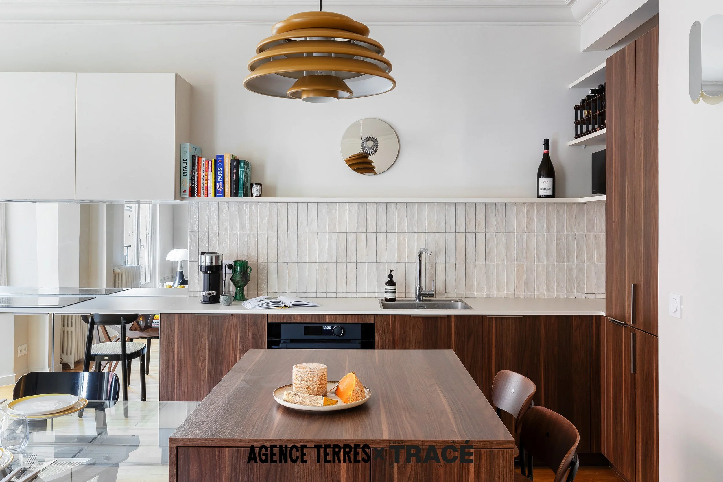 Aménagement sur mesure d'une cuisine en bois noyer avec un ilot central qui sert de bar et linéaire de cuisine en arrière plan avec un mélange de bois, de miroir et de carrelage blanc.