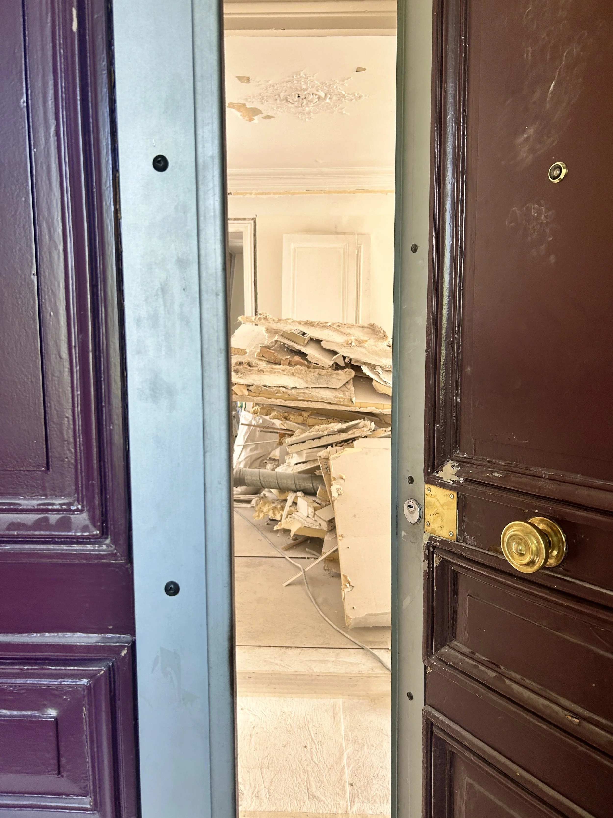 Vue à travers une porte entrouverte menant à une pièce d'un logement en cours de rénovation avec des gravats.