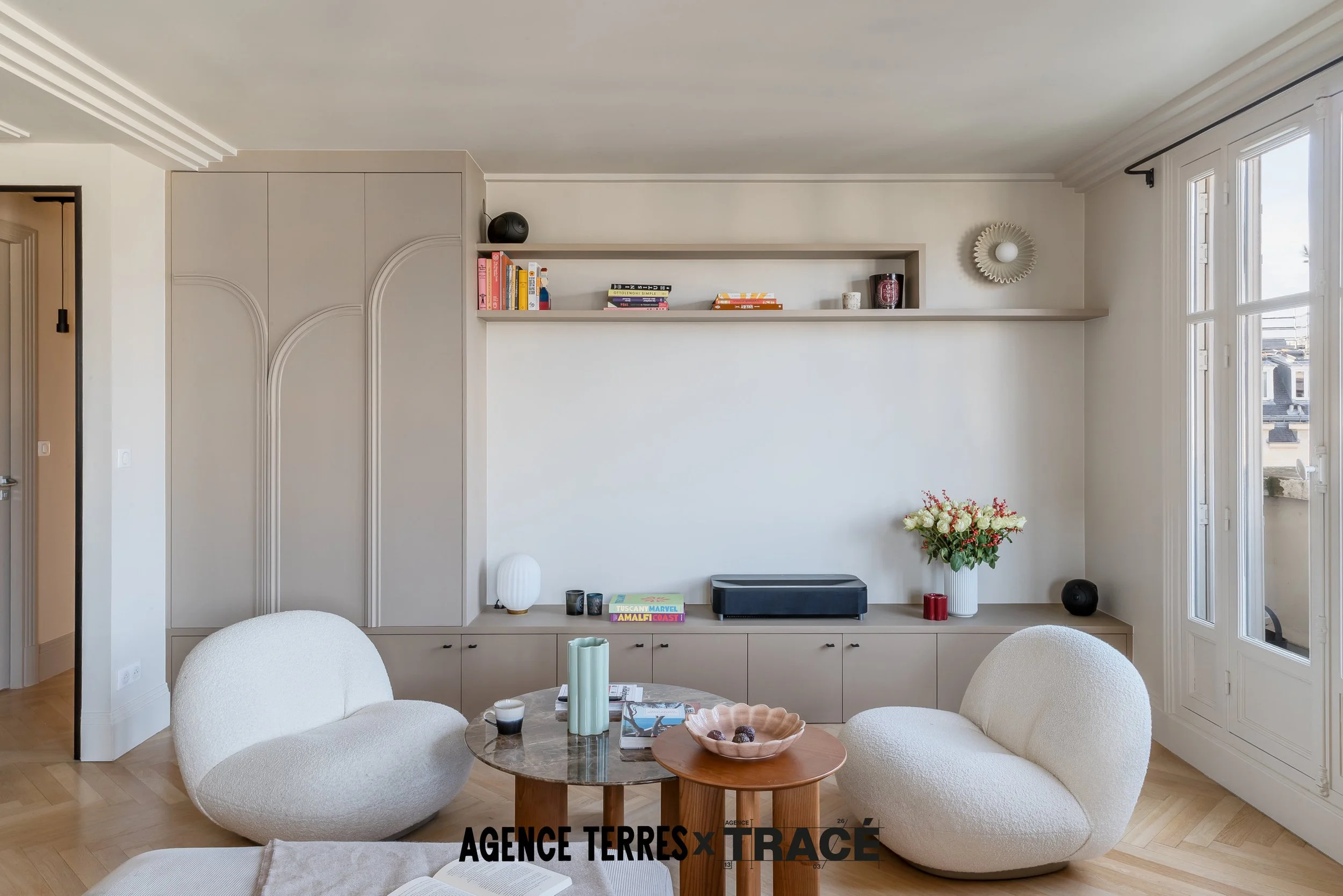 Vue d'un salon aménagé avec une bibliothèque multi fonctions avec des parties fermées pour ranger le bazar, des étagères ouvertes et un espace pour le vidéoprojecteur. Le salon est dégagé, bien aménagé dans les tons doux, lumineux et chaleureux