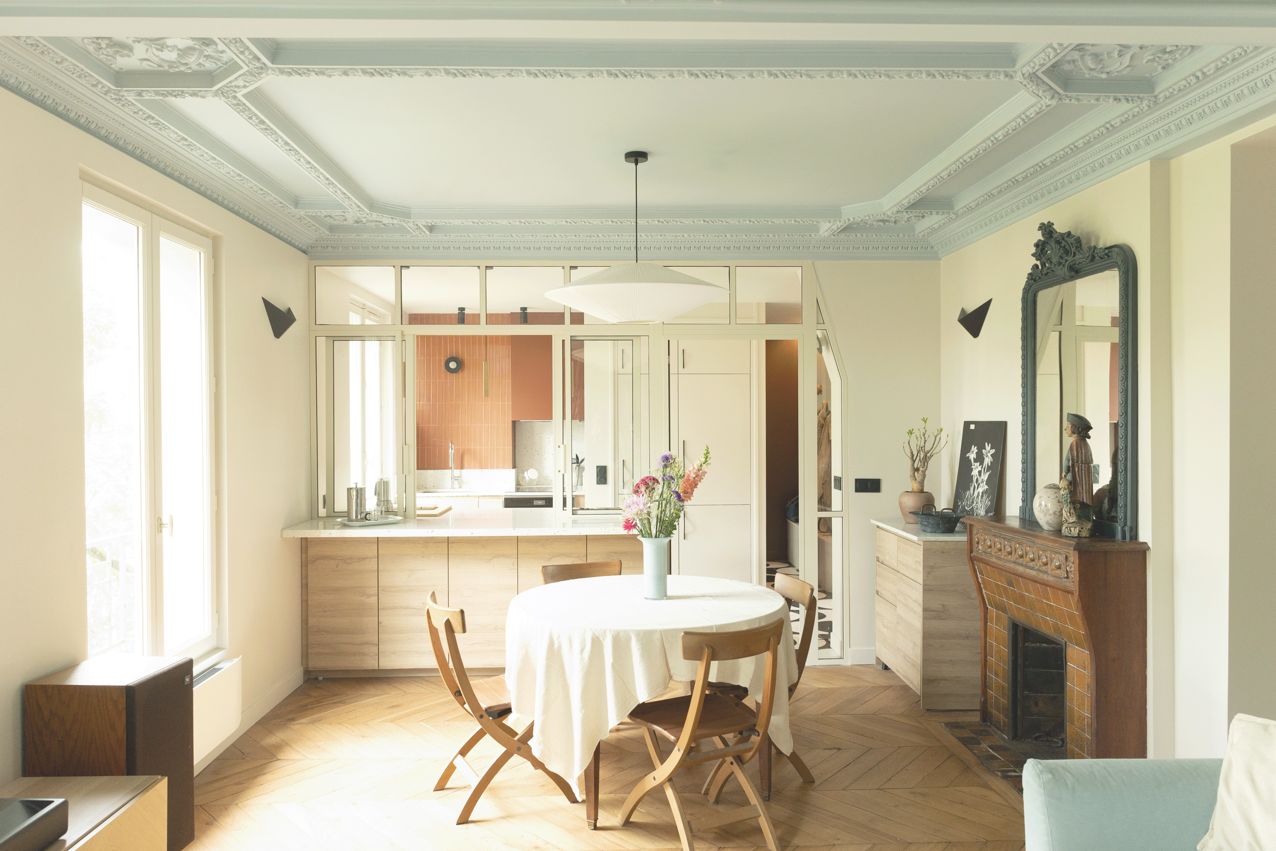 Intérieur d'une salle à manger rénovée et redécorée avec une cuisine en fond derrière une verrière vitrée sur mesure et une salle à manger en premier plan avec un parquet restauré, de nouvelles peintures et notamment un plafond bleu.