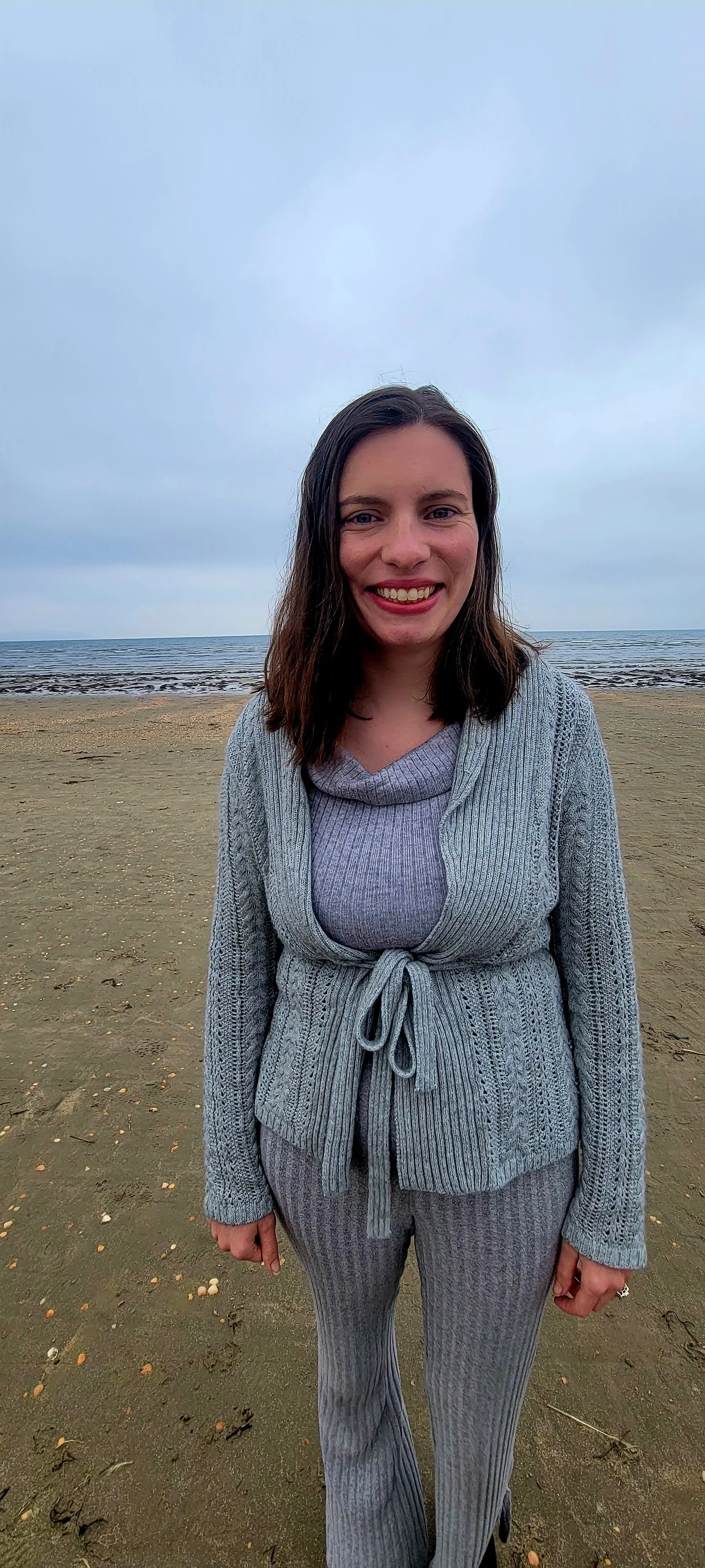 En kvinna som ler på en strand under en molnig himmel, klädd i en grå stickad kofta som är knuten i midjan, en lila polotröja och randiga byxor.