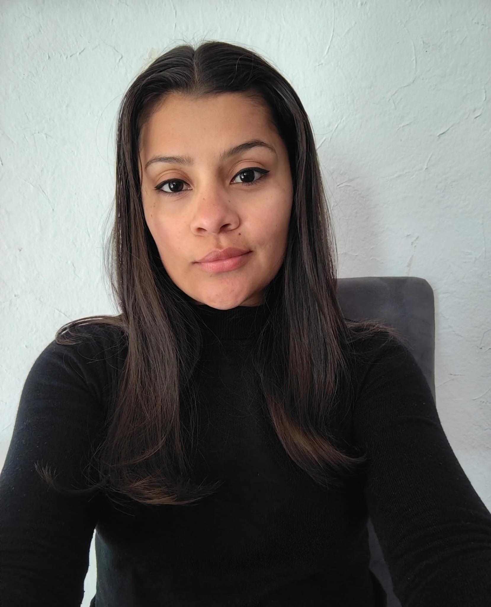 Retrato de una mujer con cabello liso y oscuro, usando maquillaje con delineador, con fondo de pared blanca.
