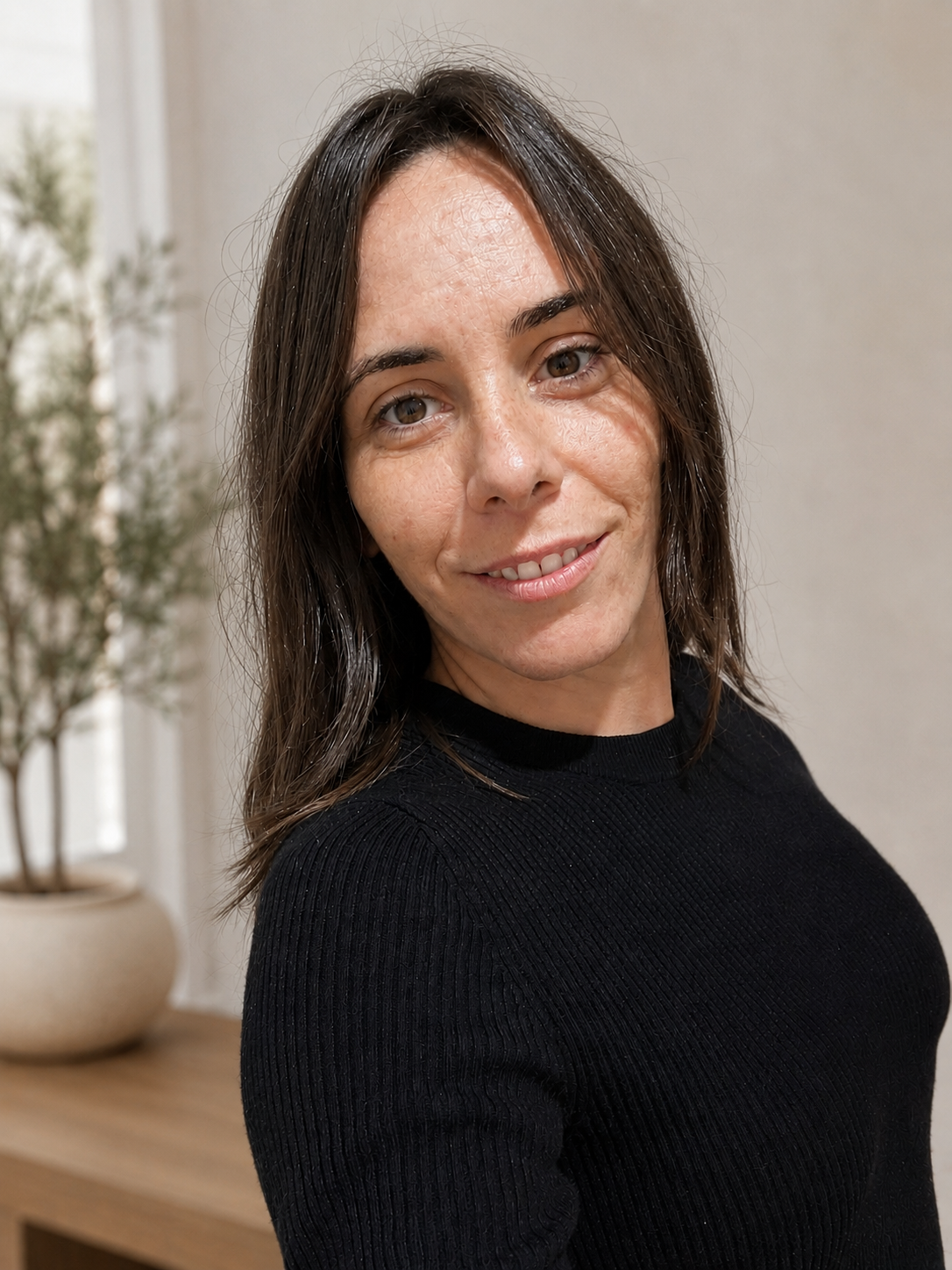 Retrato de una mujer con cabello castaño oscuro, piel clara y sonrisa, usando un suéter negro, en un ambiente interior con planta en el fondo.