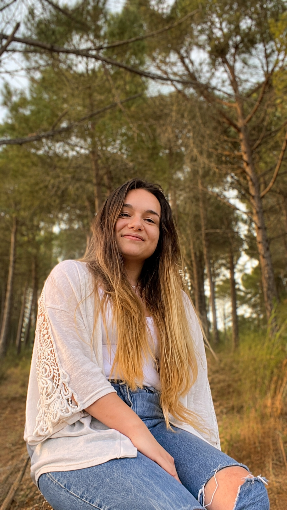 Joven mujer sonriendo, sentada en un bosque de pinos, con ropa blanca y pantalones azules con roto, cabello largo y castaño claro.