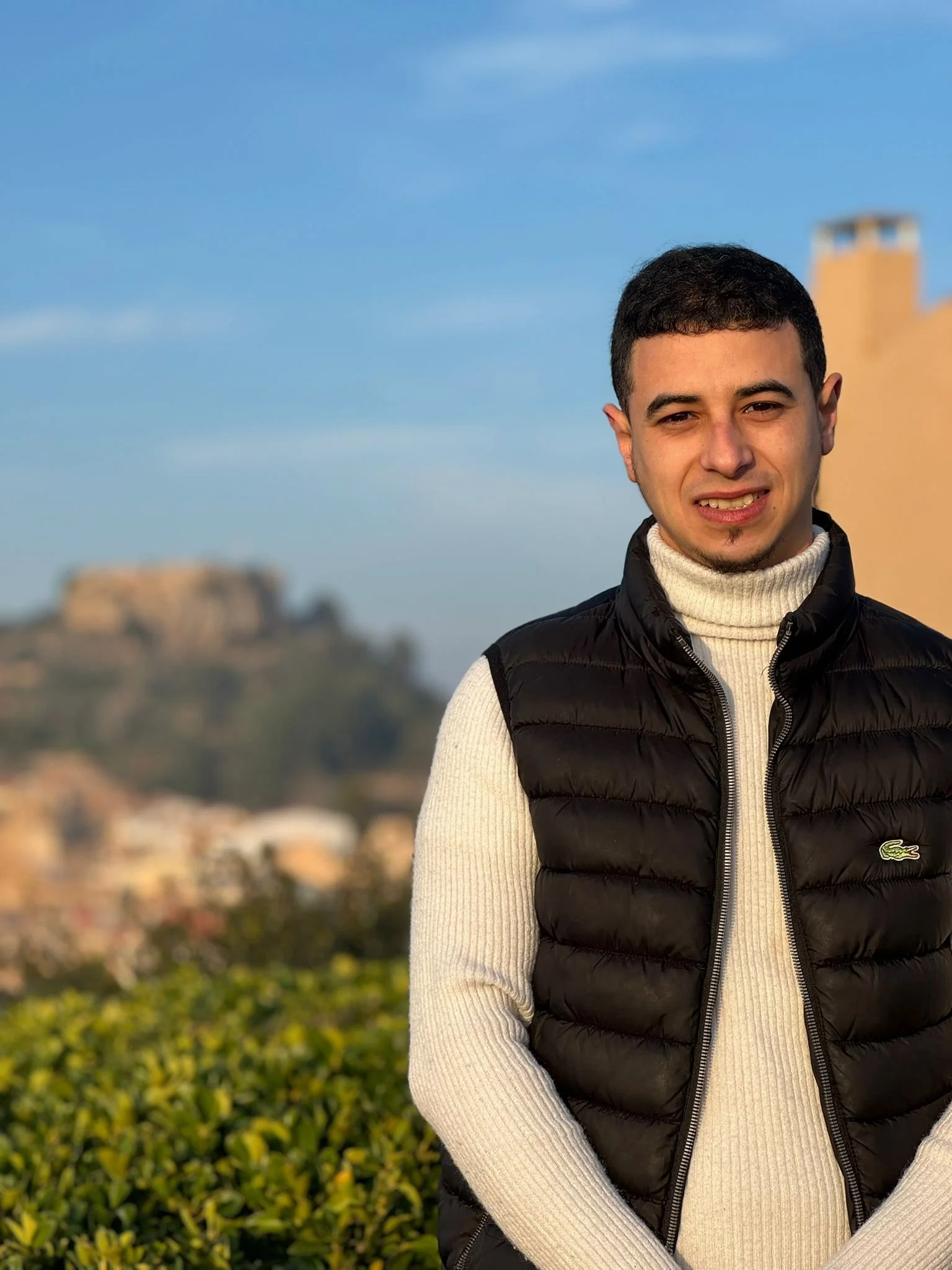 Joven hombre con piel clara, cabello oscuro y corto, viste un suéter de color crema y un chaleco negro con logo de Lacoste, posando al aire libre con fondo de paisaje urbano y cielo despejado.