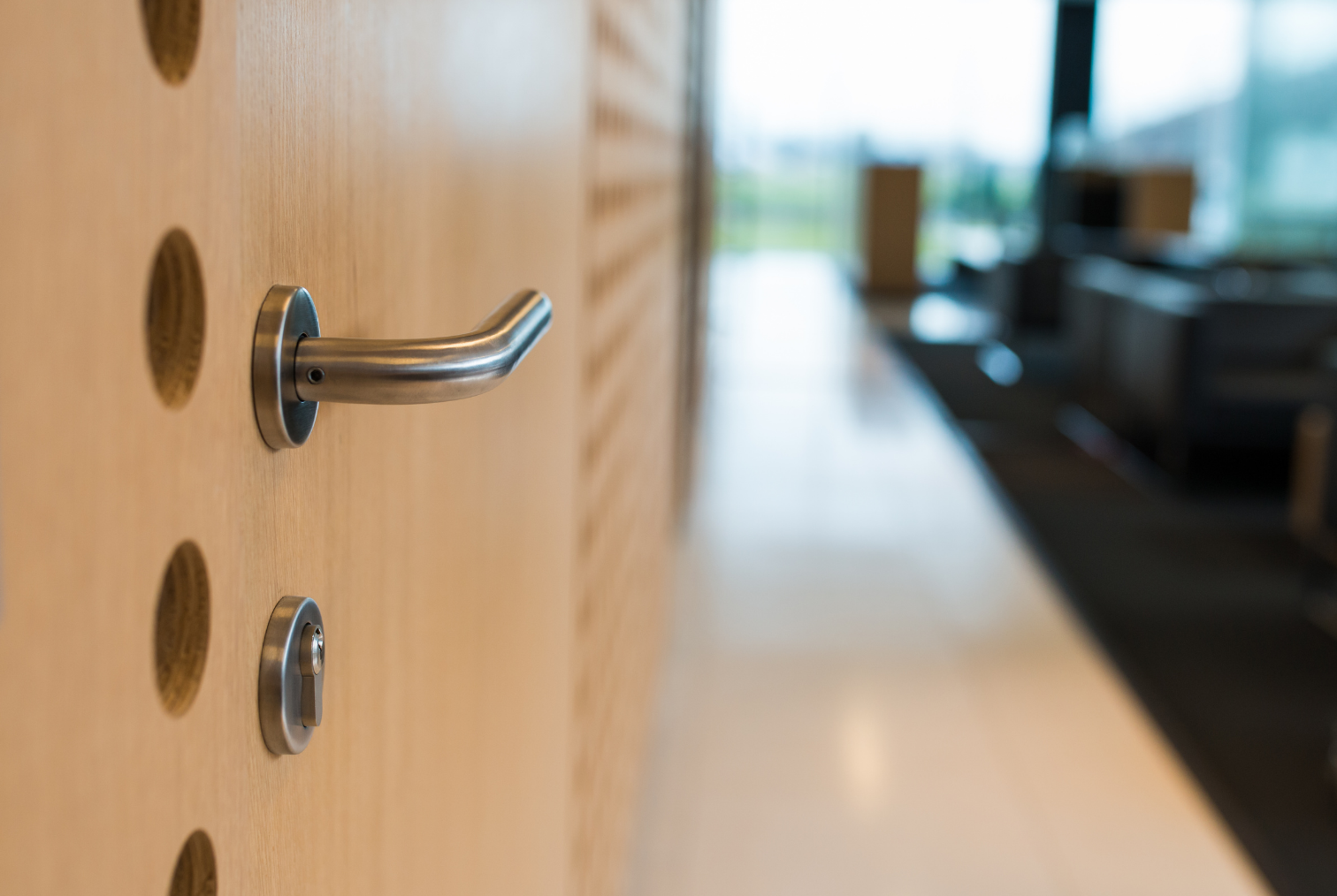 Poignée de porte en métal fixé à une surface en bois clair avec des trous circulaires. L'arrière-plan montre un intérieur moderne avec des meubles et de grandes fenêtres donnant sur un extérieur vert.
