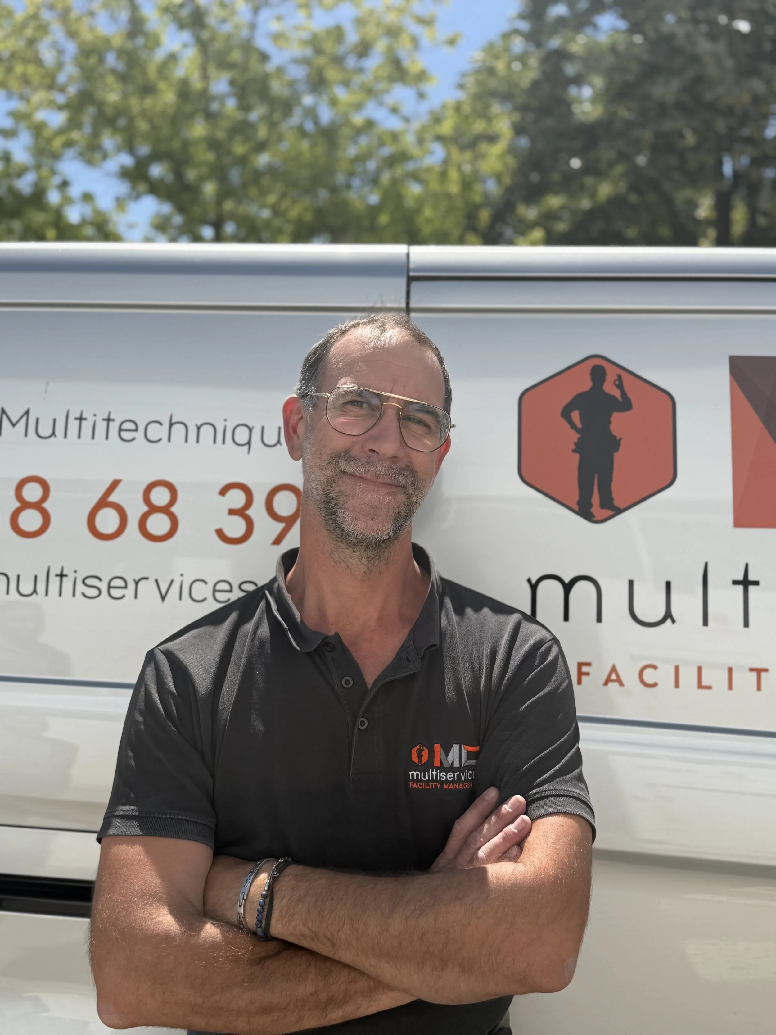 Un homme souriant portant une chemise noire avec un logo multitechnique, avec des bras croisés, devant un camion blanc avec un logo et des textes en français, sous un ciel bleu avec des arbres en arrière-plan.