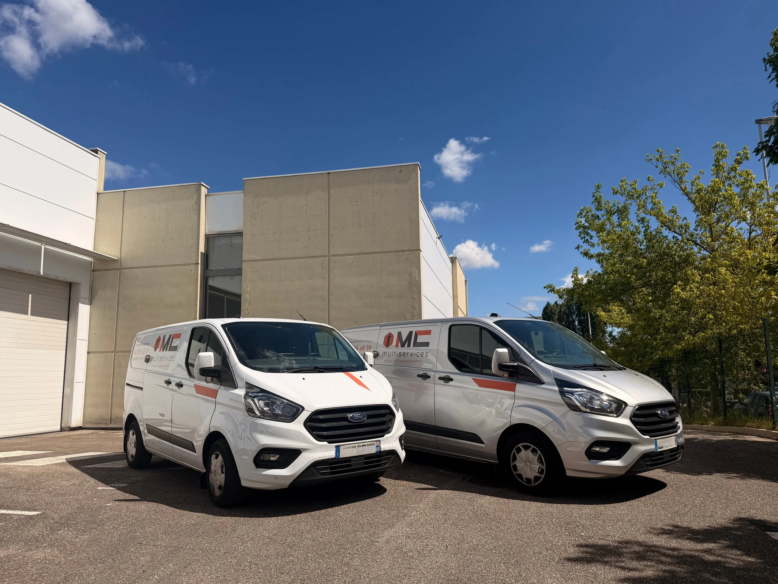 Vans blancs garés devant un bâtiment moderne avec mur beige, ciel bleu clair avec quelques nuages, et arbres verts à droite.
