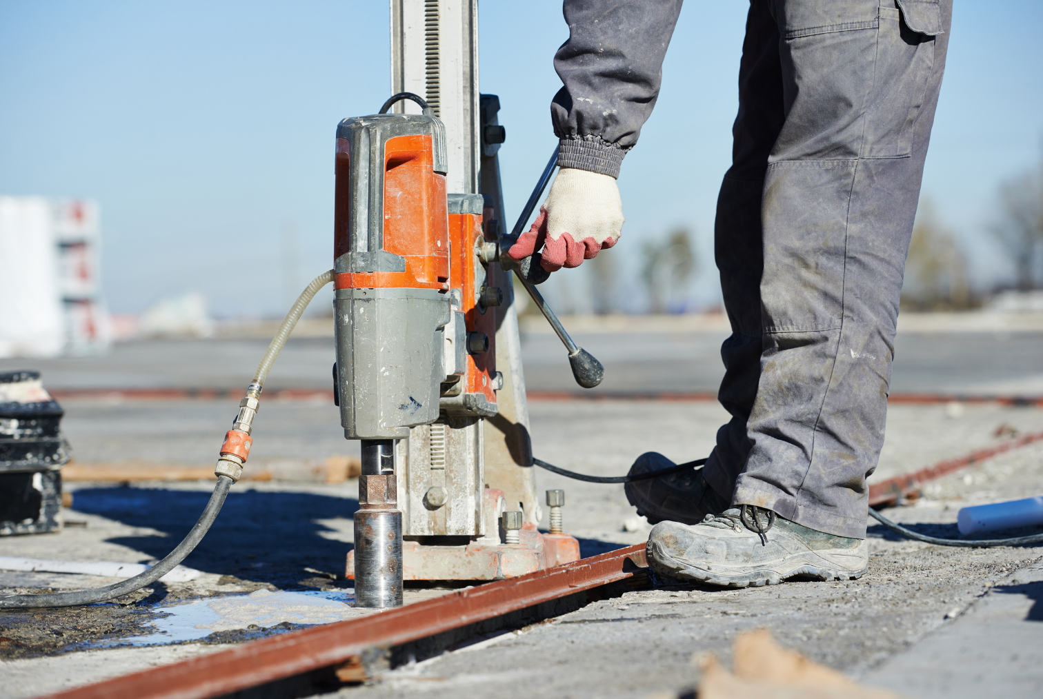 Travailleur utilisant une machine de forage sur un chantier de construction, portant des vêtements de travail, des gants et des chaussures renforcées, en extérieur sous un ciel clair.