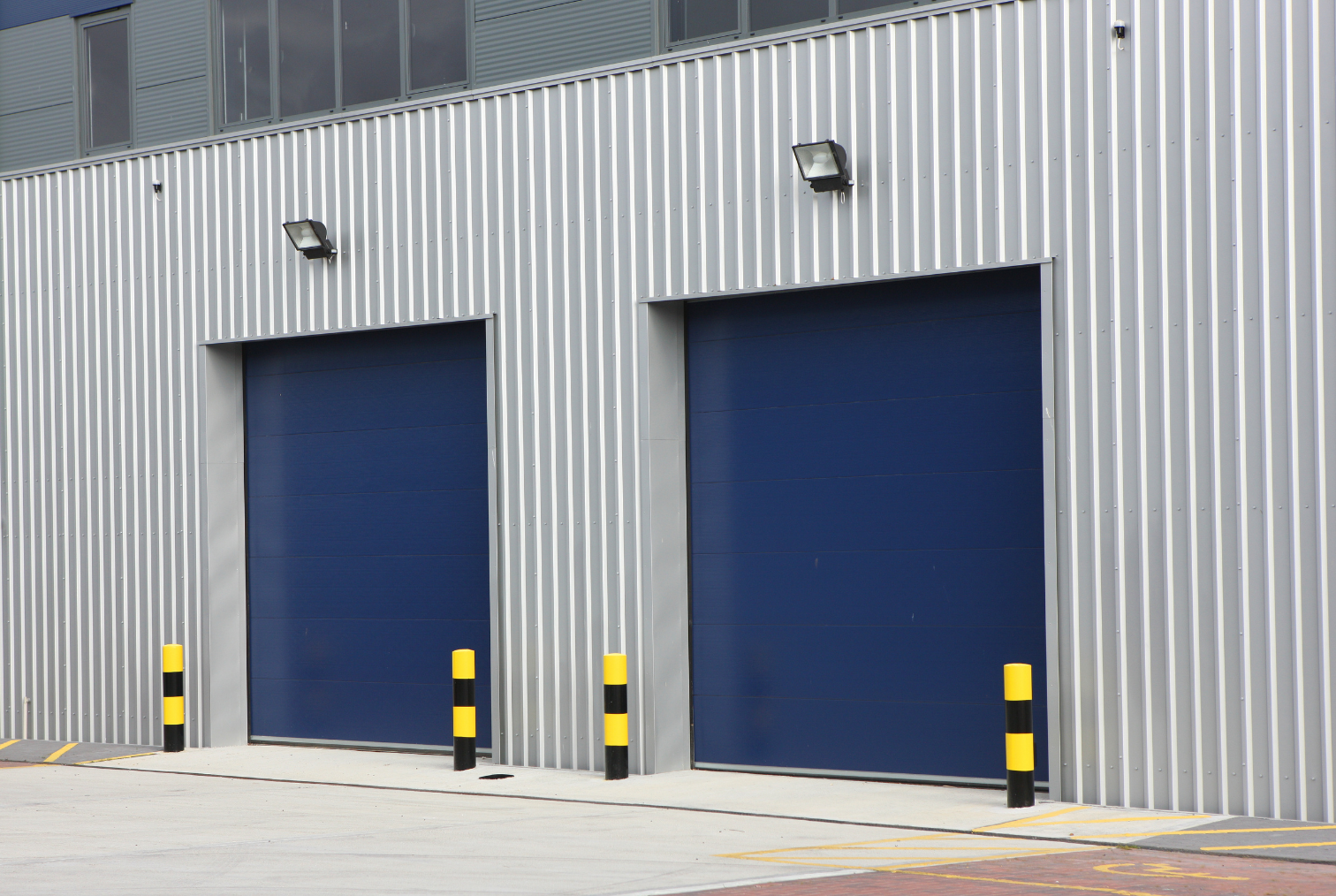 Deux portes de garage bleues sur un bâtiment en métal avec un éclairage et des bornes anti-intrusion jaunes et noires à l'avant.