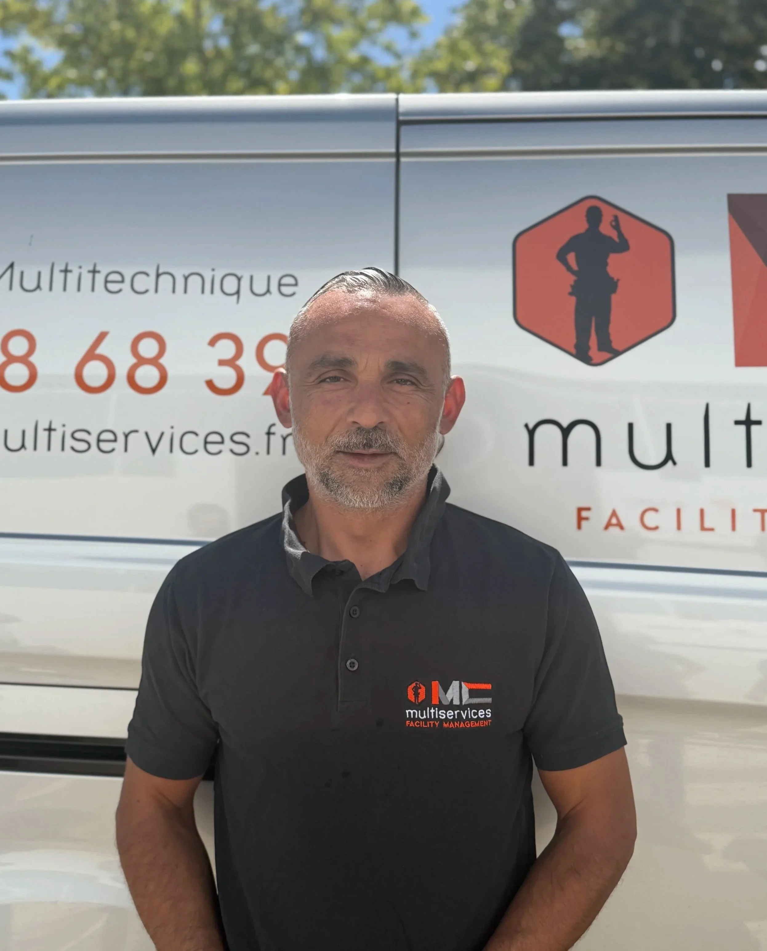 Homme portant un t-shirt noir avec le logo de MultiServices Facility Management devant un véhicule blanc avec des icônes et du texte en français.
