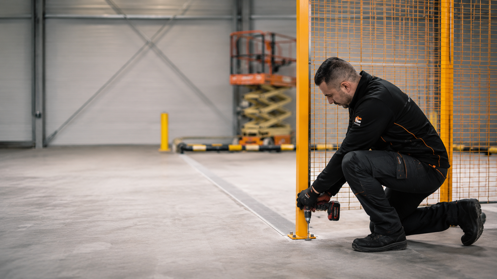 Un homme en uniforme de travail utilisant une perceuse pour fixer une barrière métallique orange au sol dans un entrepôt ou un espace industriel.