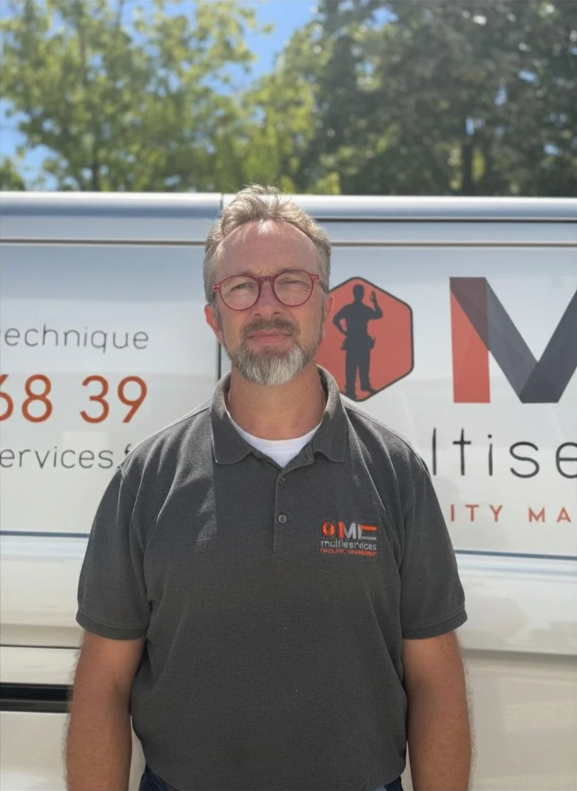 Un homme avec des lunettes et une barbe regarde l'appareil photo devant un véhicule avec une signalisation d'entreprise de services techniques.