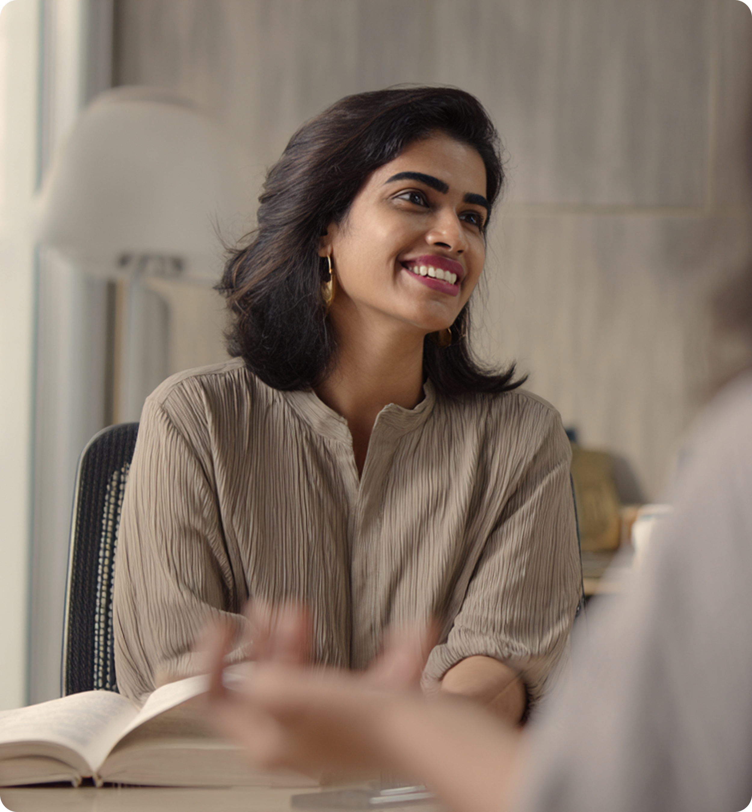 A woman with dark hair, dark eyebrows, and hoop earrings smiling while looking to the right. She wears a brown striped shirt and is sitting at a desk with an open notebook in front of her in a professional setting.