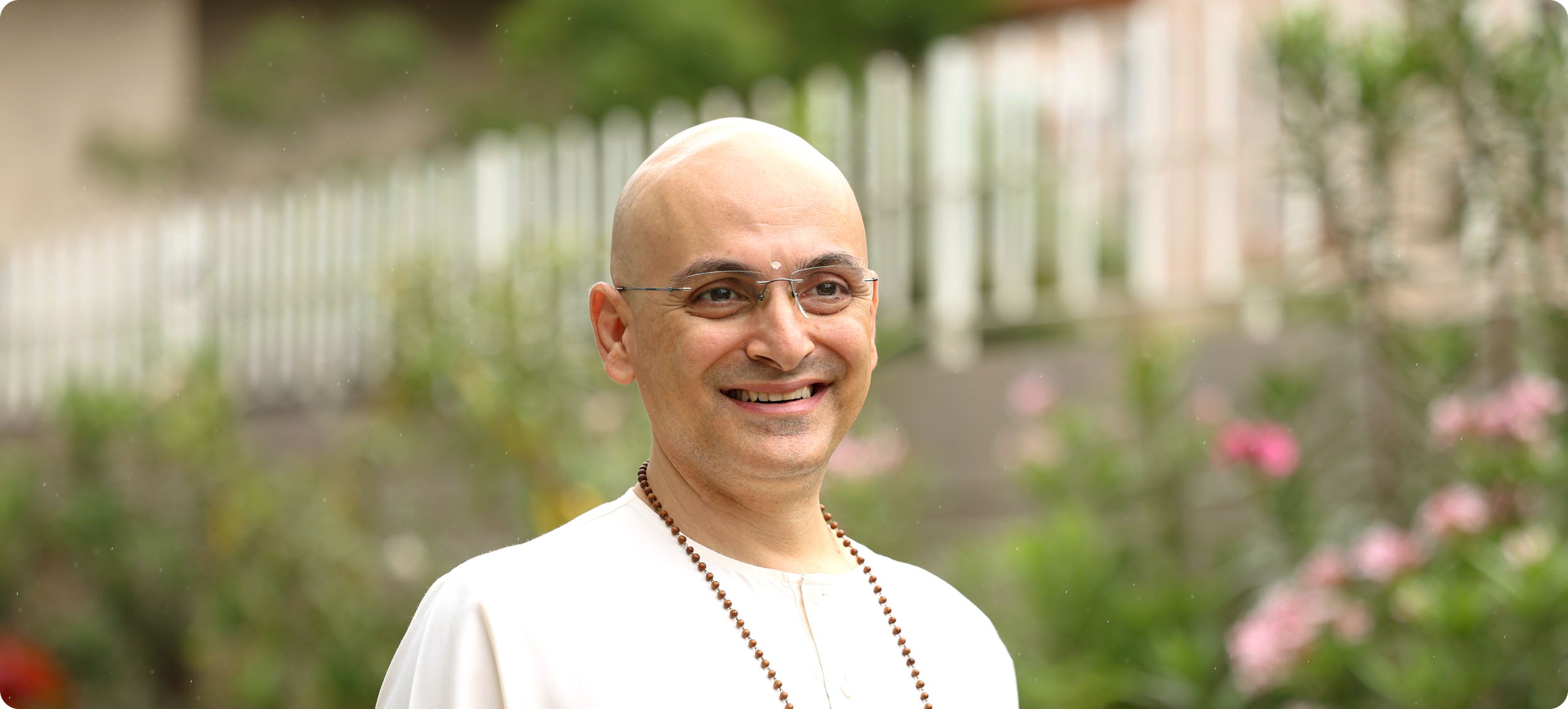 A smiling man with a shaved head wearing glasses and a white shirt outdoors, with a fence, greenery, and pink flowers in the background.