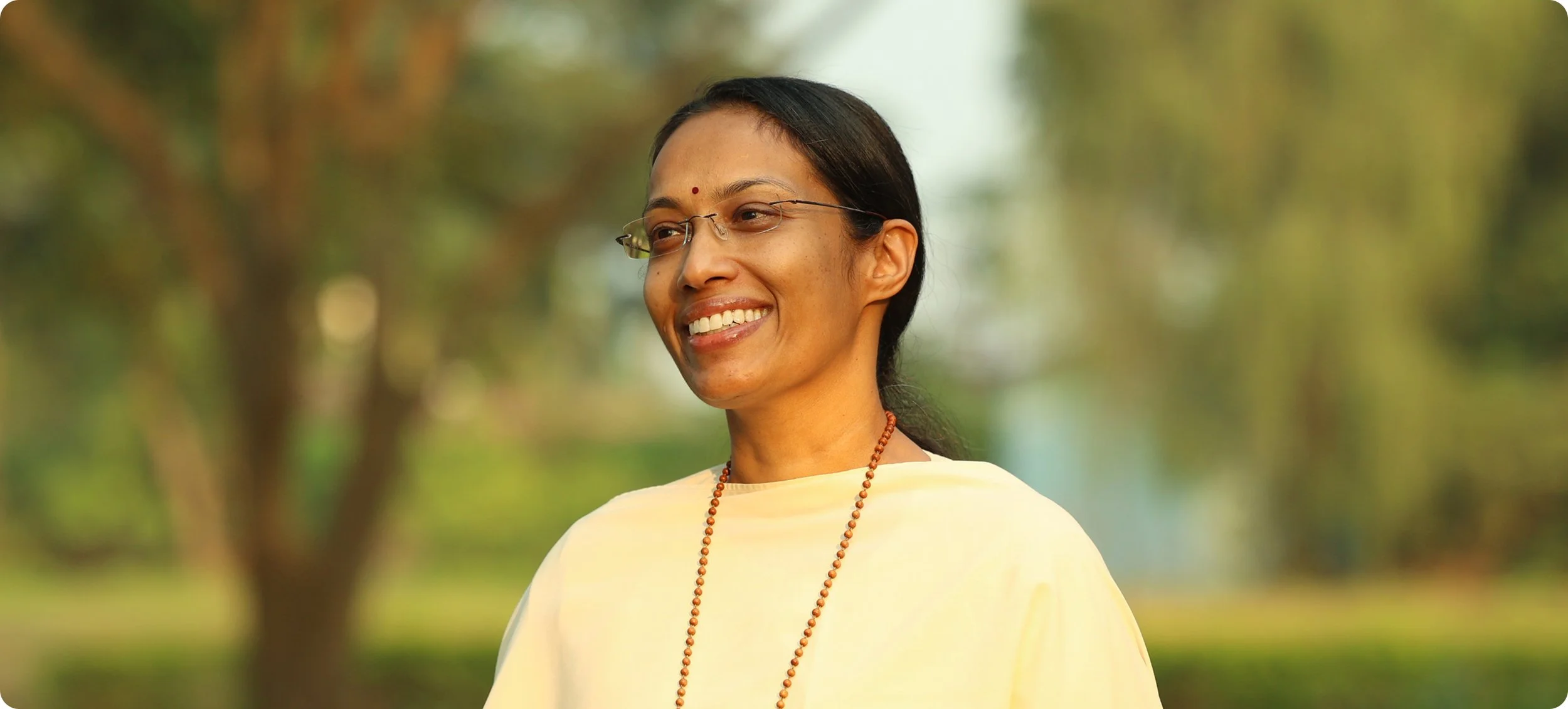 Smiling woman wearing glasses and a yellow outfit outdoors with a blurred green and brown background.