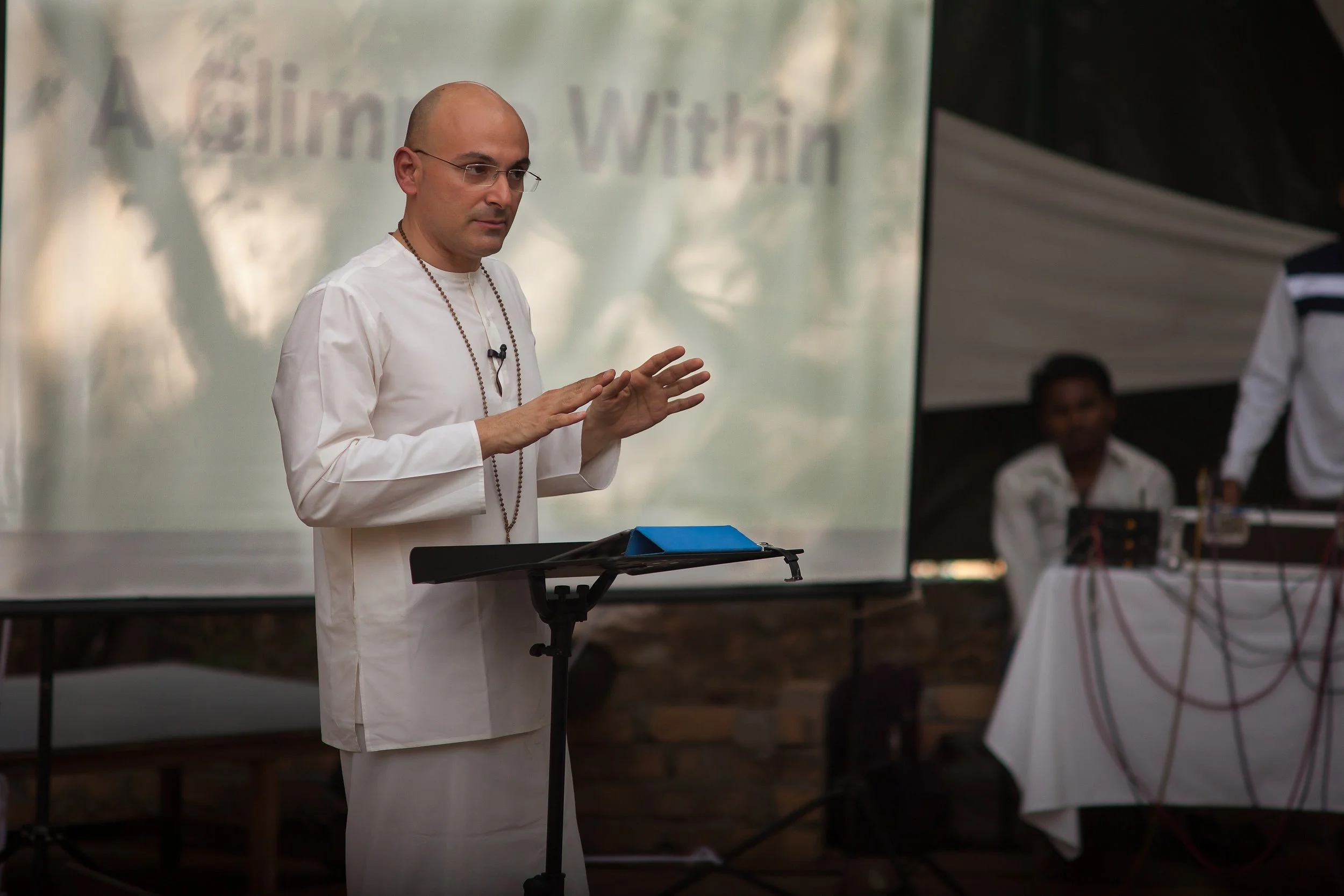 A man wearing a white traditional outfit, glasses, and prayer beads, standing at a podium with a tablet, speaking in front of a large screen that shows part of the word 'Achieving' and 'Within'.