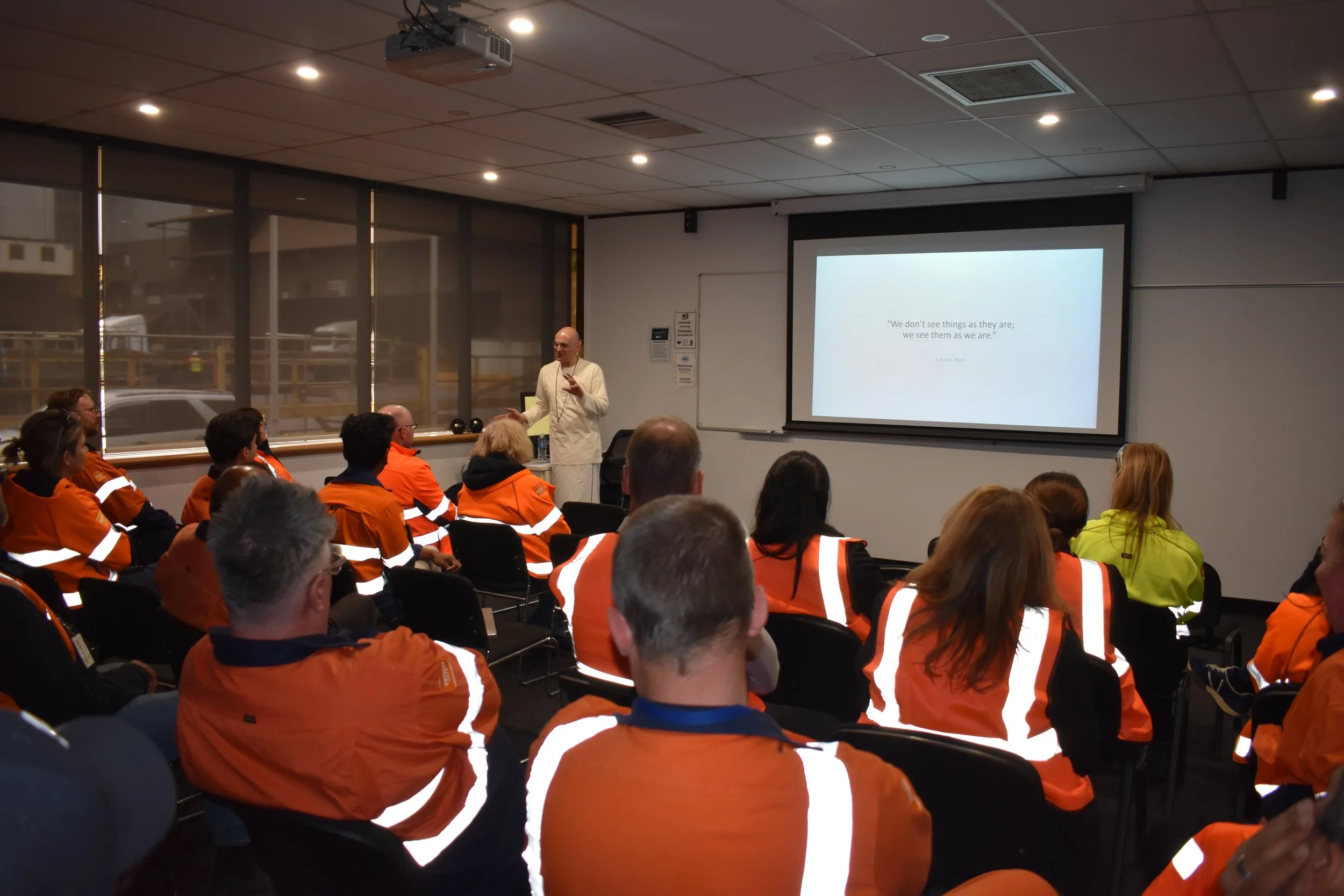 A man in white traditional attire is standing and speaking in front of a seated audience of people in orange safety jackets. A presentation slide on the screen behind him displays a quote about perception.