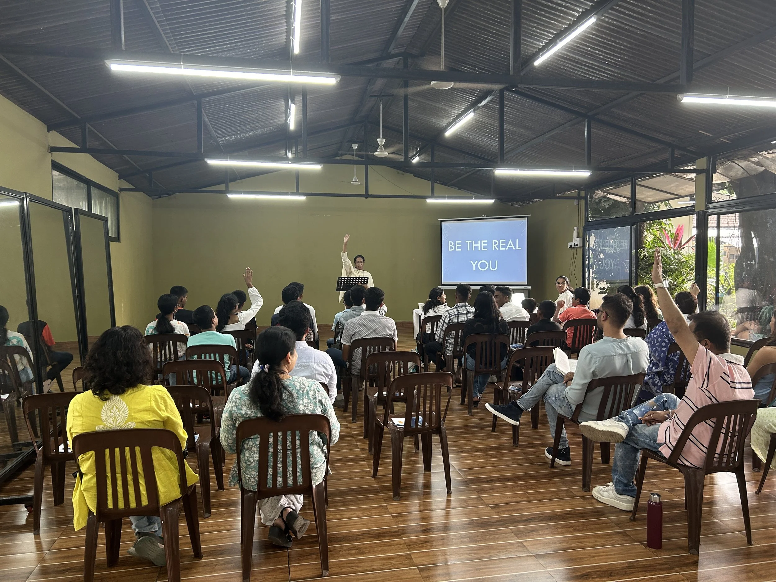 A classroom or seminar room with many people seated and raising their hands. A woman is standing at the front, speaking into a microphone. A screen behind shows the words 'BE THE REAL YOU'. The room has large windows with greenery outside, high ceiling with exposed metal roof, and ceiling fans.