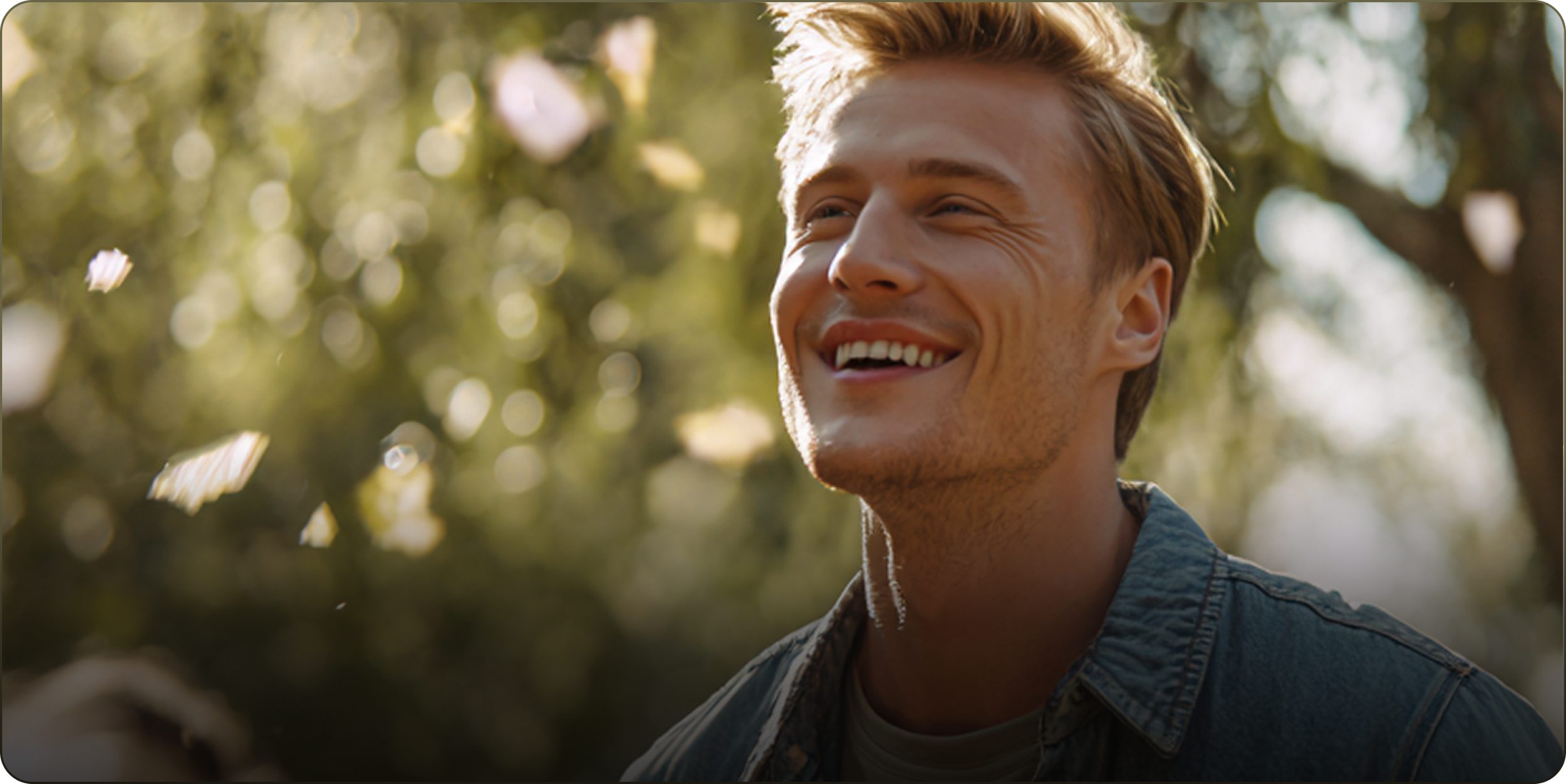 A young man with blonde hair smiling happily outdoors among trees with sunlight filtering through the leaves.