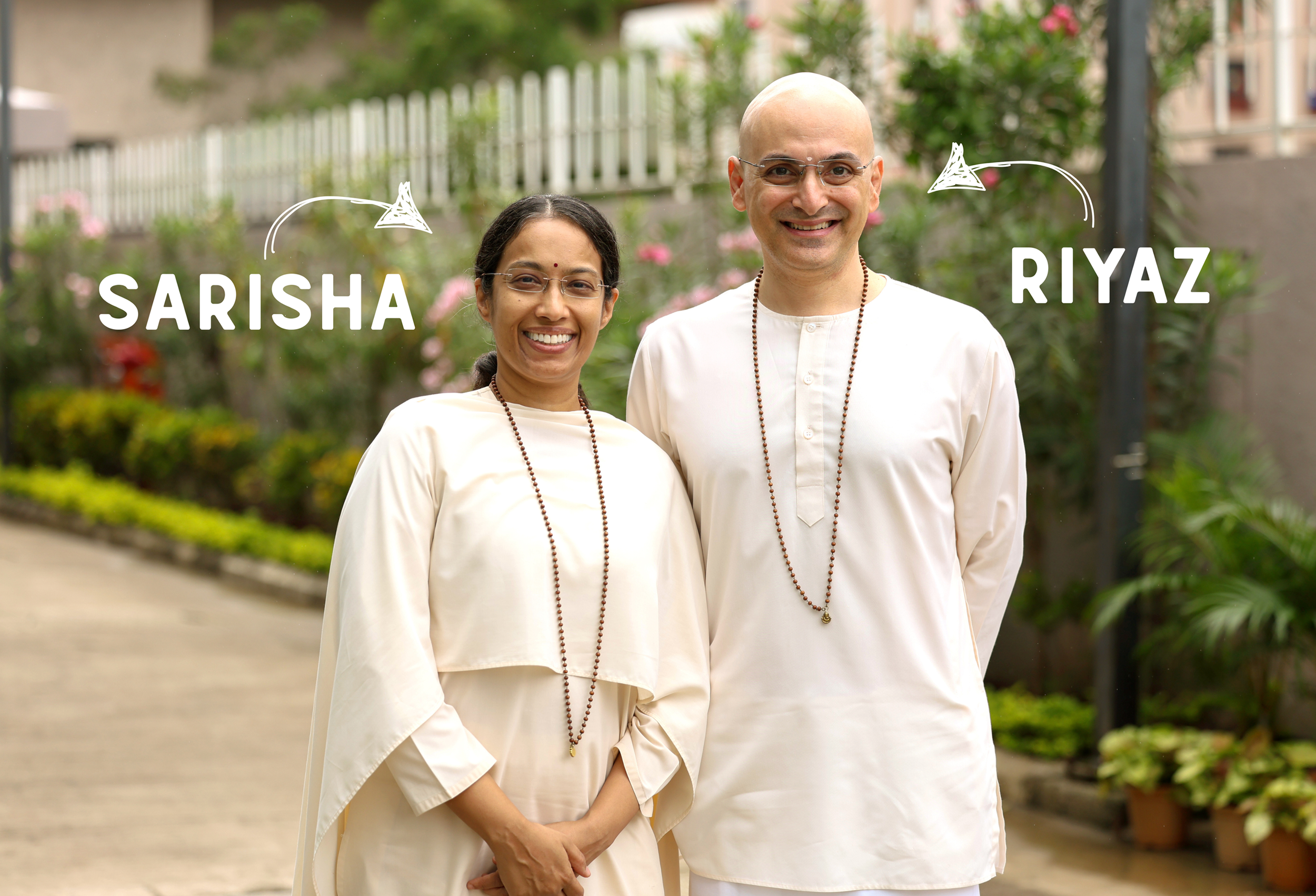 A smiling woman and man standing outdoors in front of a garden with pink flowers and a white fence. The woman has glasses and is wearing a cream-colored outfit with a beaded necklace. The man is bald, wearing glasses and a cream-colored kurta with a beaded necklace.