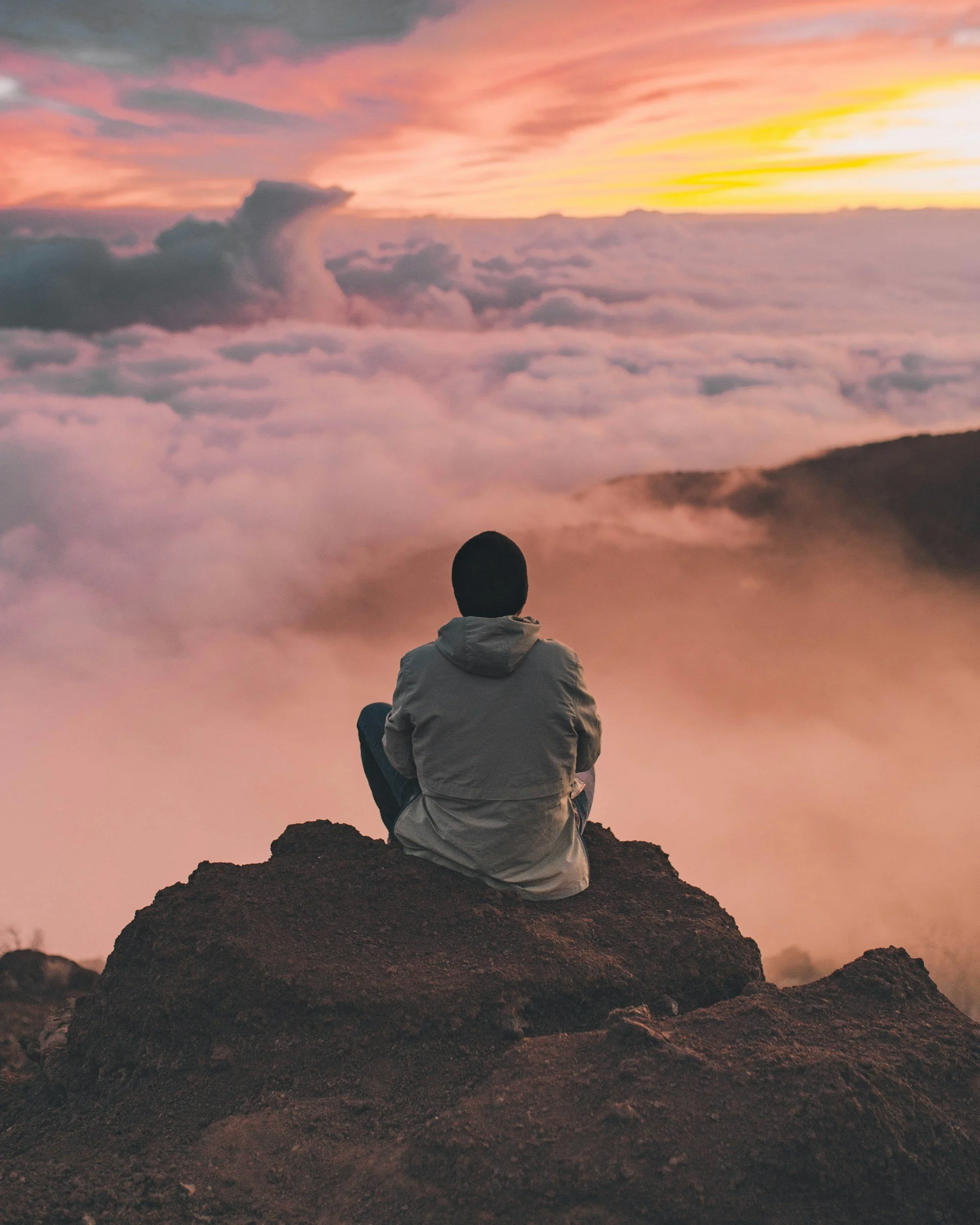 Person sitting on rocky terrain gazing at the colorful sunset sky with clouds.