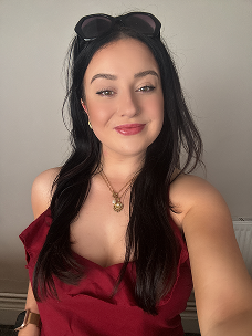 A young woman with long dark hair wearing a red top, sunglasses on her head, and a gold necklace, posing indoors against a plain gray wall.